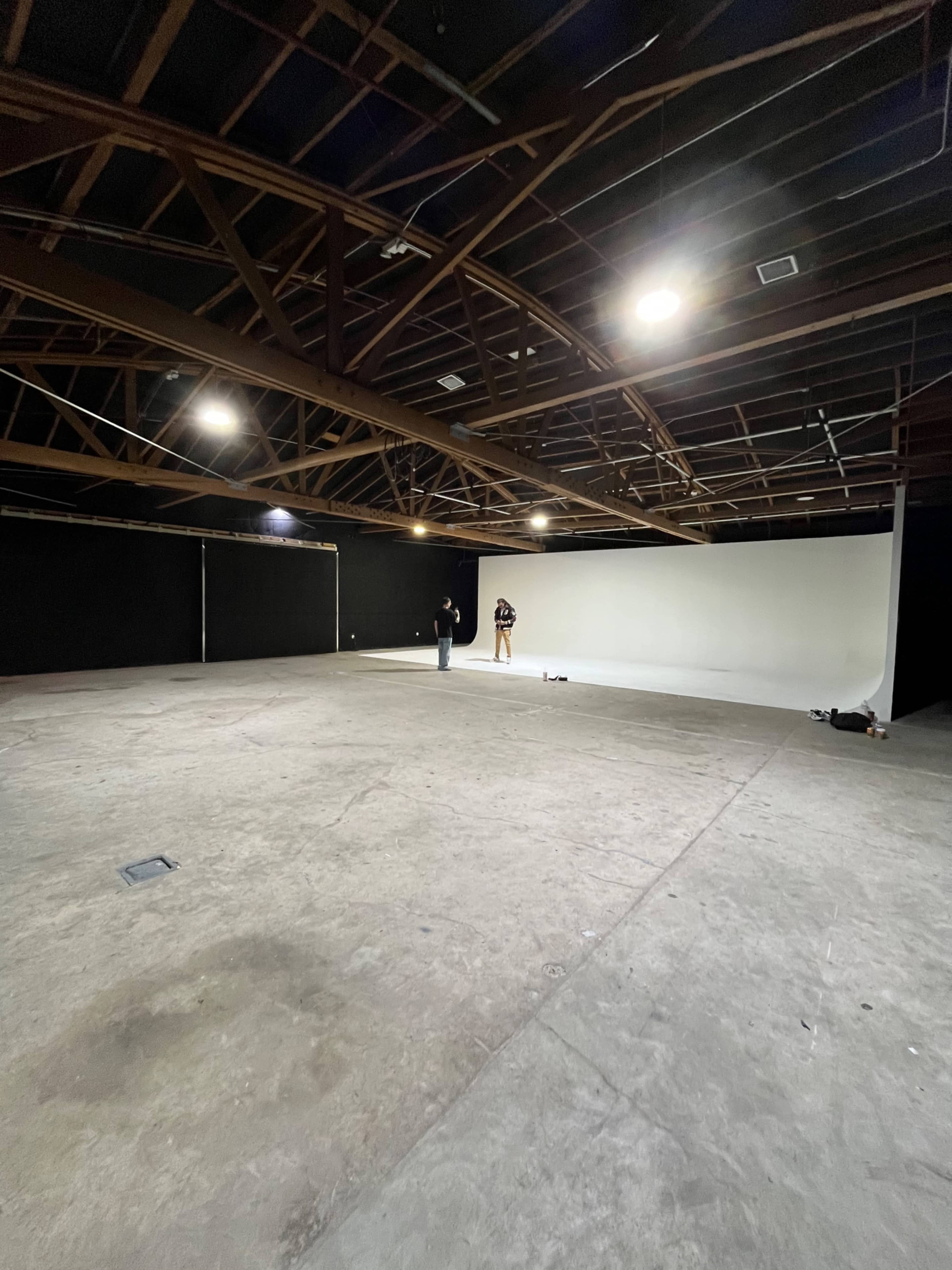 The image shows a large, empty studio space with a concrete floor, a white backdrop at one end, and two individuals positioned within the room.