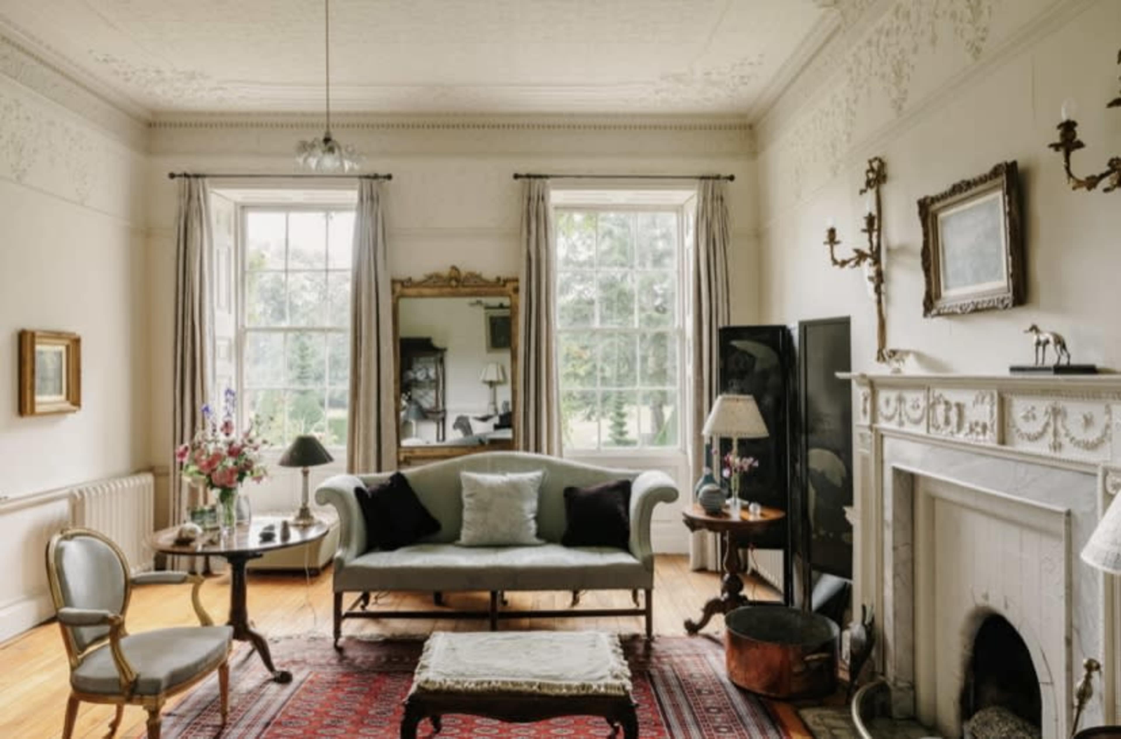 The room features a sofa, a round table, a large mirror, and a fireplace, all set against a backdrop of windows with curtains and a patterned rug on the wooden floor.