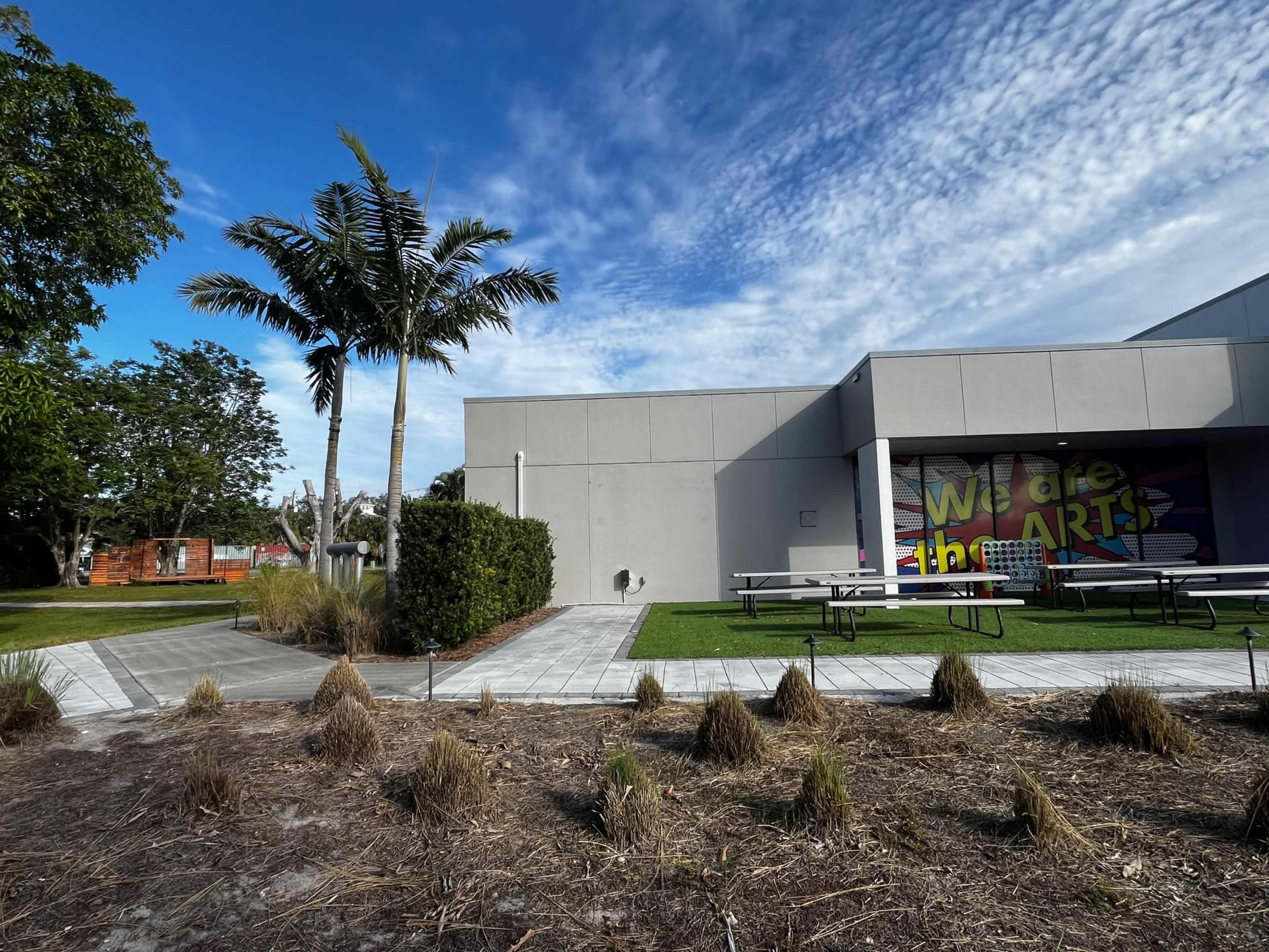 A building with colorful wall art and picnic tables is surrounded by landscaped greenery and palm trees under a partly cloudy sky.