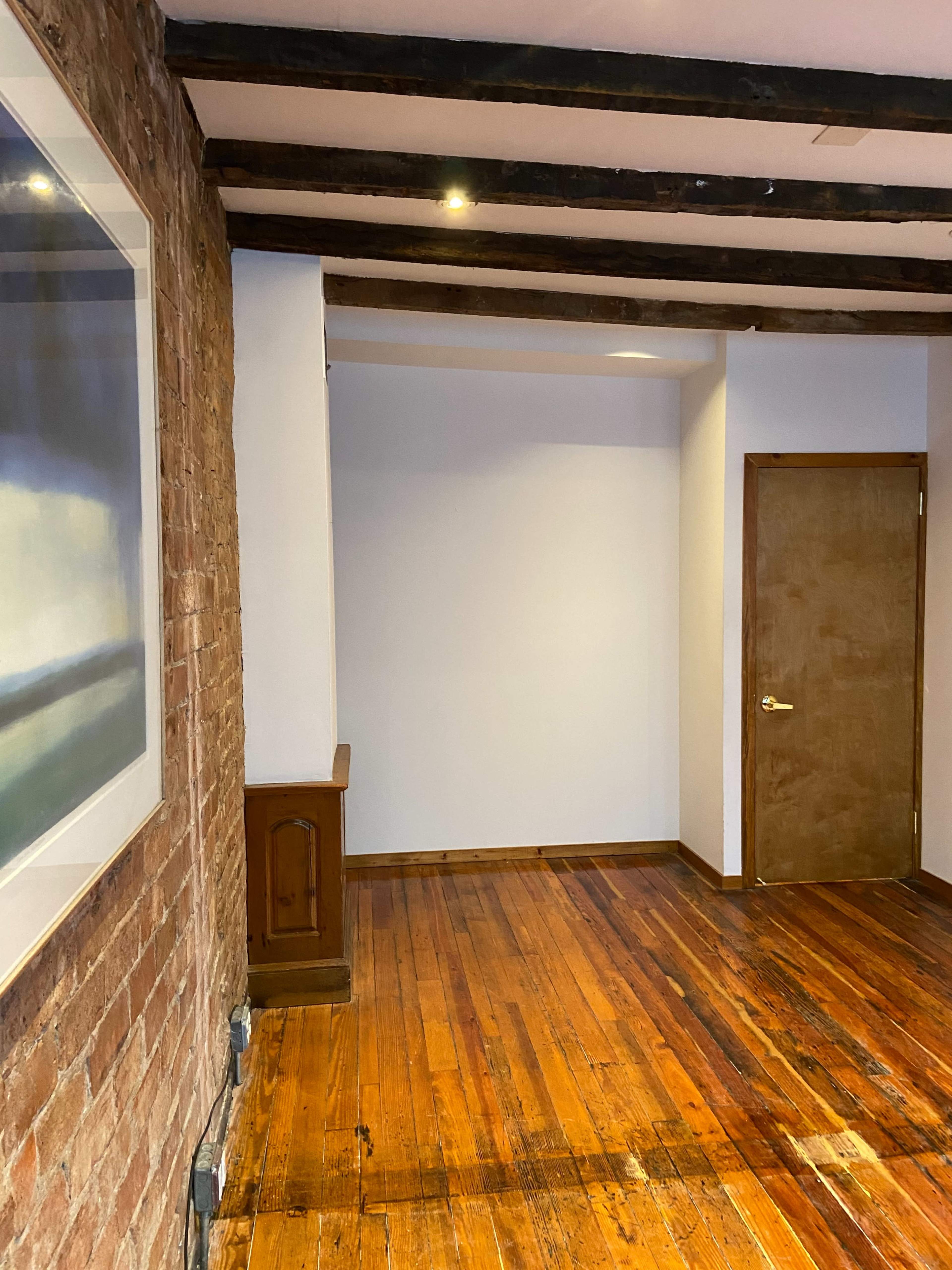 A room featuring exposed wooden beams, a brick wall, and hardwood floors with a plain white wall and a closed wooden door in the corner.