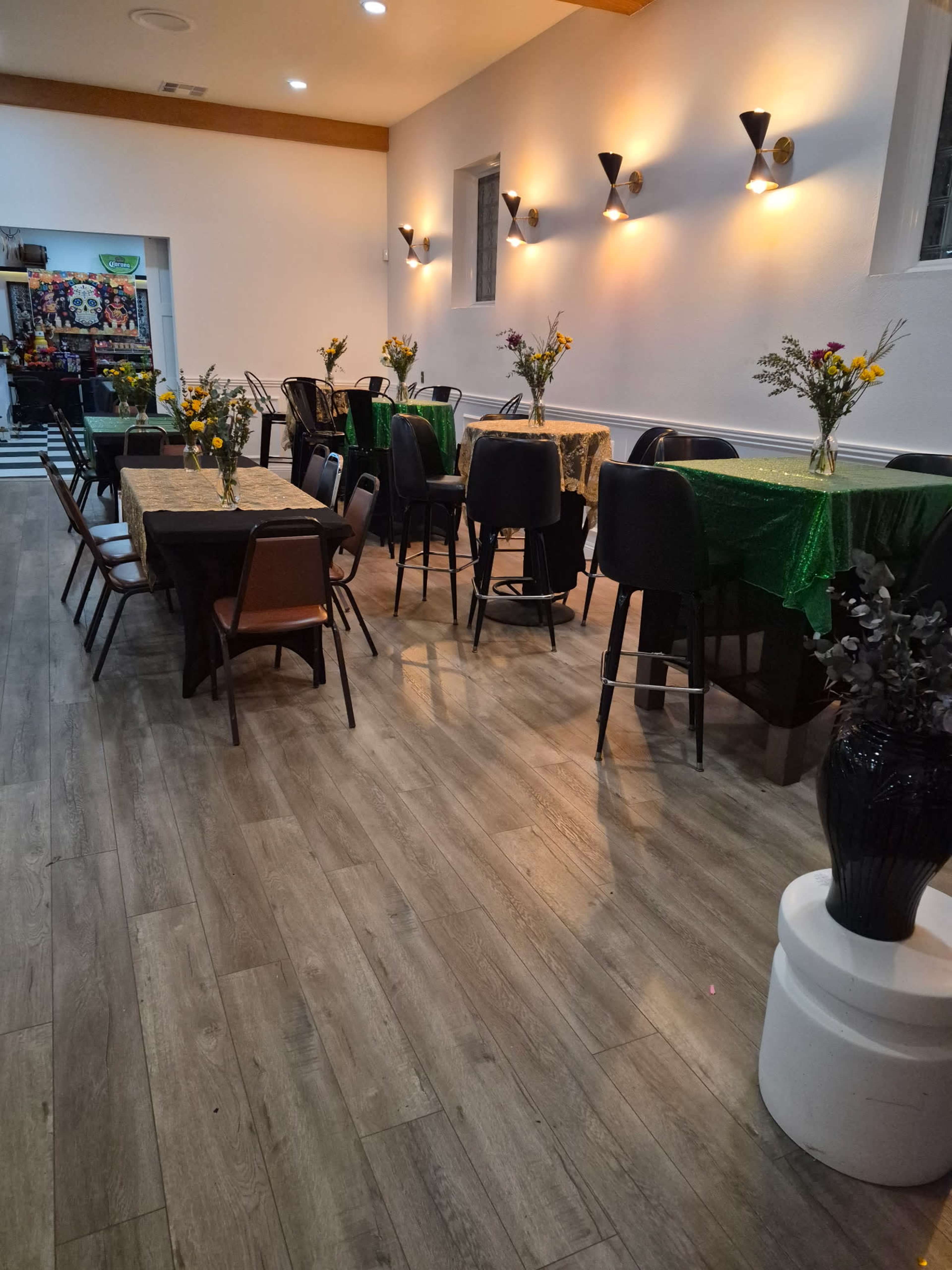The image shows a dining area with tables covered in colorful cloths, chairs arranged around them, and decorative floral centerpieces, illuminated by wall-mounted lighting.