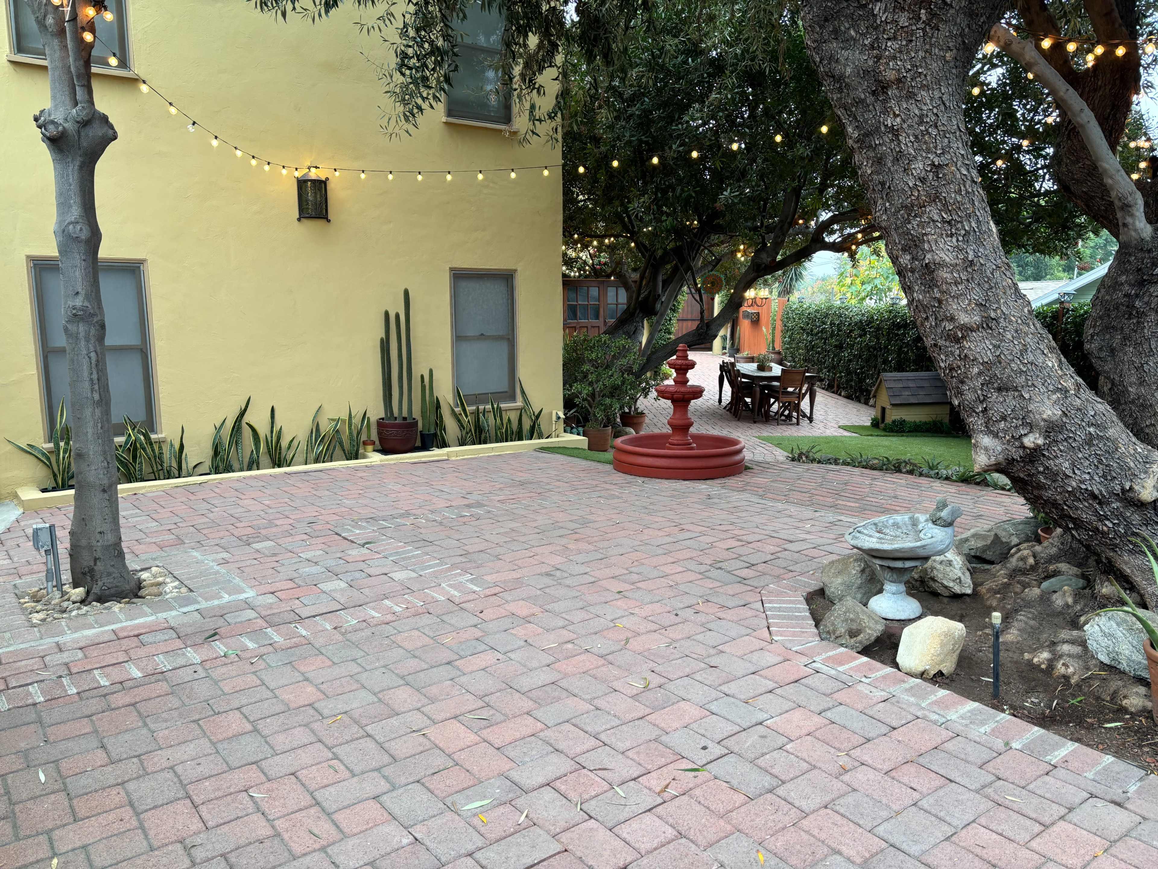 The scene features a brick-paved courtyard with a red fountain, surrounding greenery, and string lights overhead.