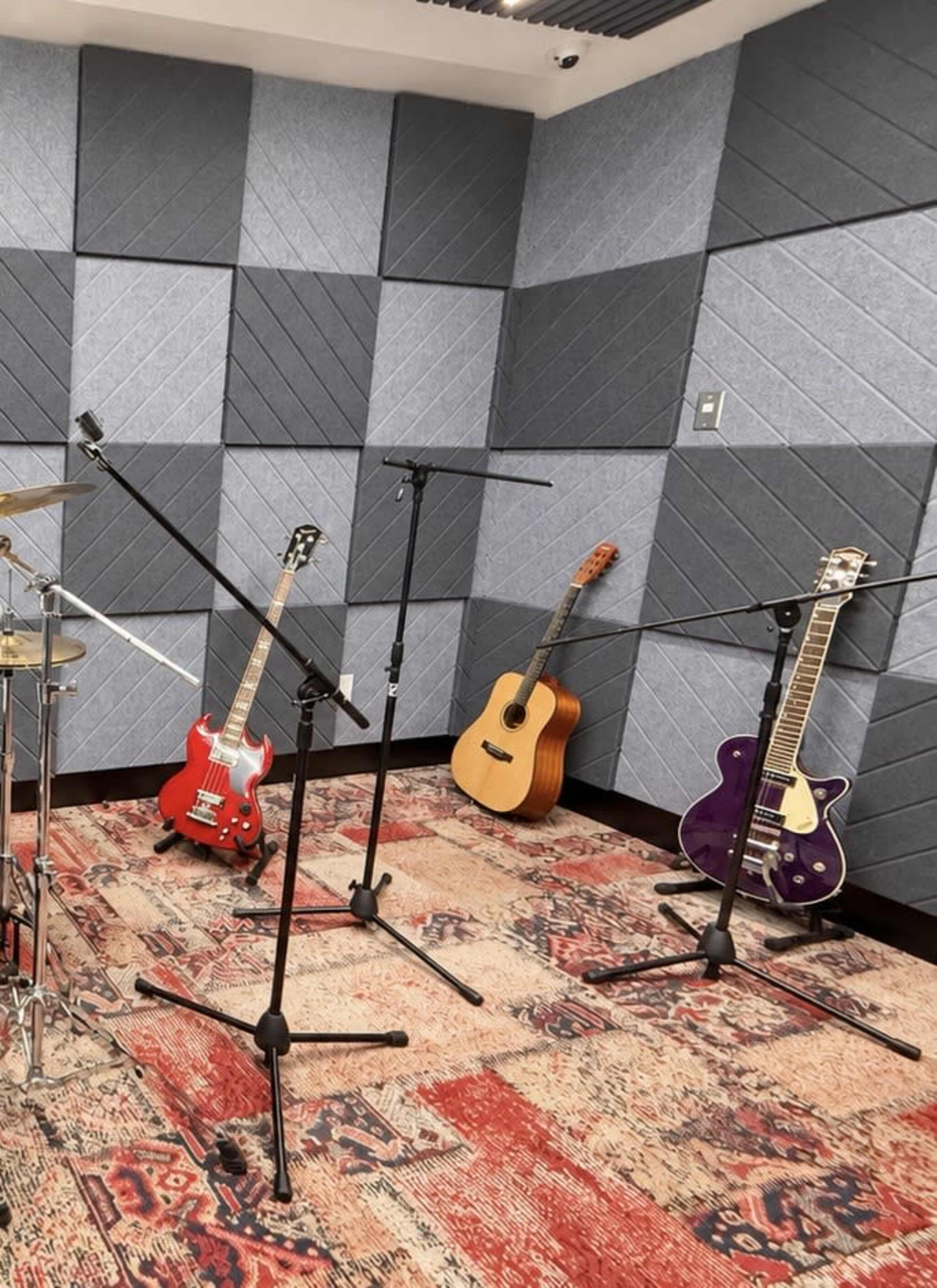 The image shows a music studio with various musical instruments, including a red electric guitar, a purple electric guitar, an acoustic guitar, and a drum set, all placed on a patterned rug and surrounded by soundproof wall panels.