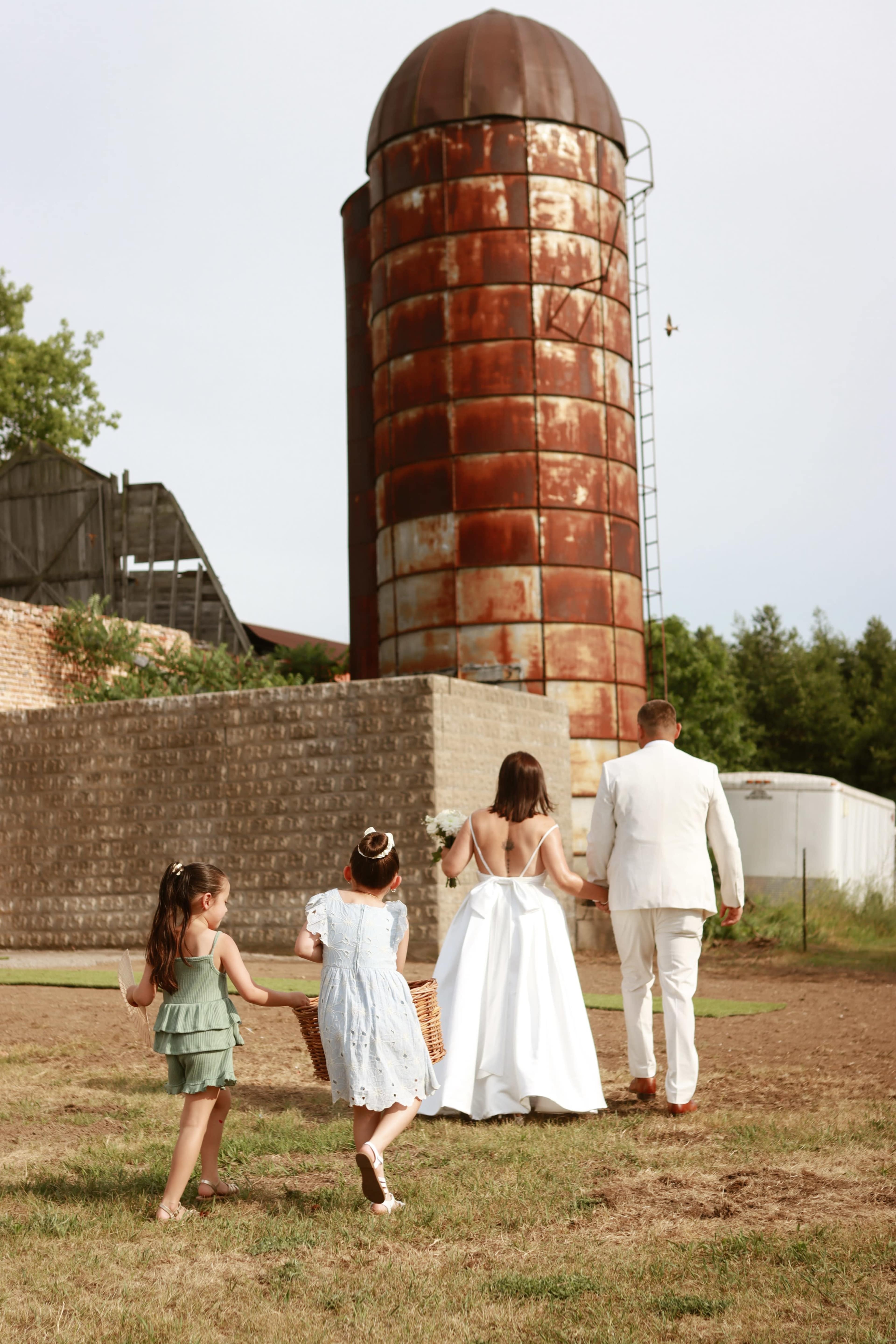 A bride and groom walk toward a weathered silo with two young girls following them, one carrying a basket.