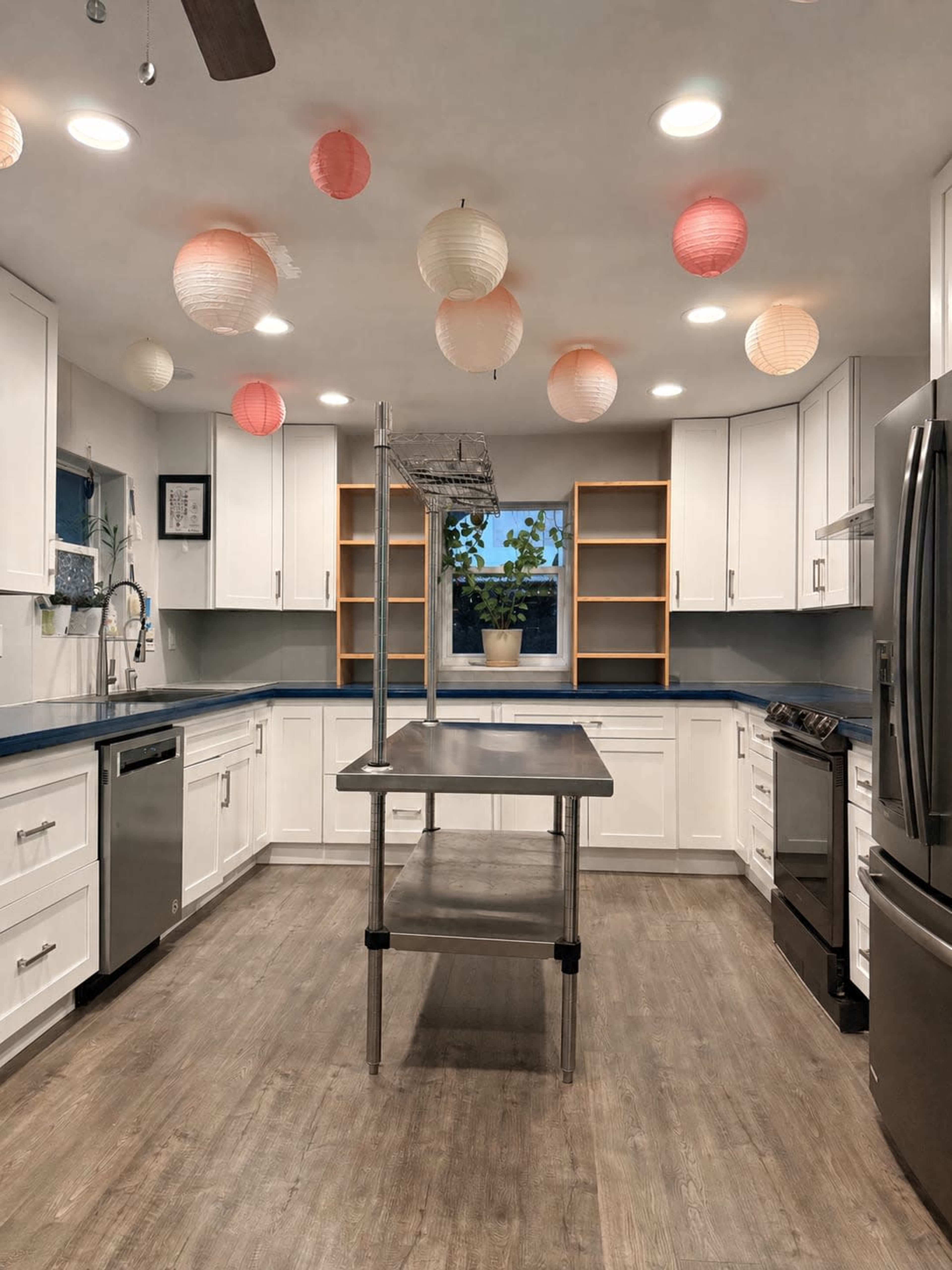 A modern kitchen features white cabinetry, stainless steel appliances, a central metal island, and paper lanterns hanging from the ceiling.