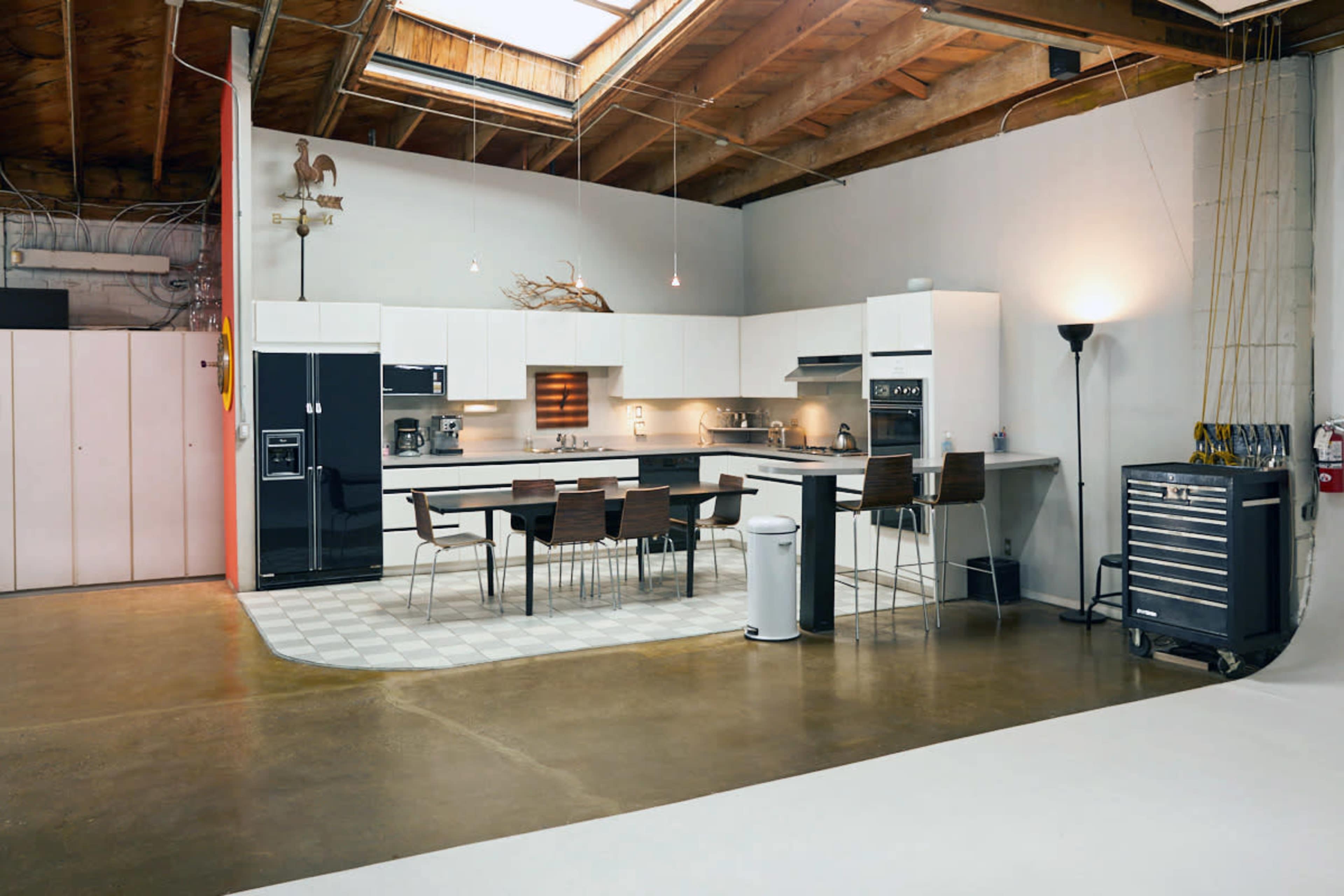 A modern kitchen features white cabinetry, a black refrigerator, and a dining table with four brown chairs, all set against a concrete floor and exposed wooden beams.