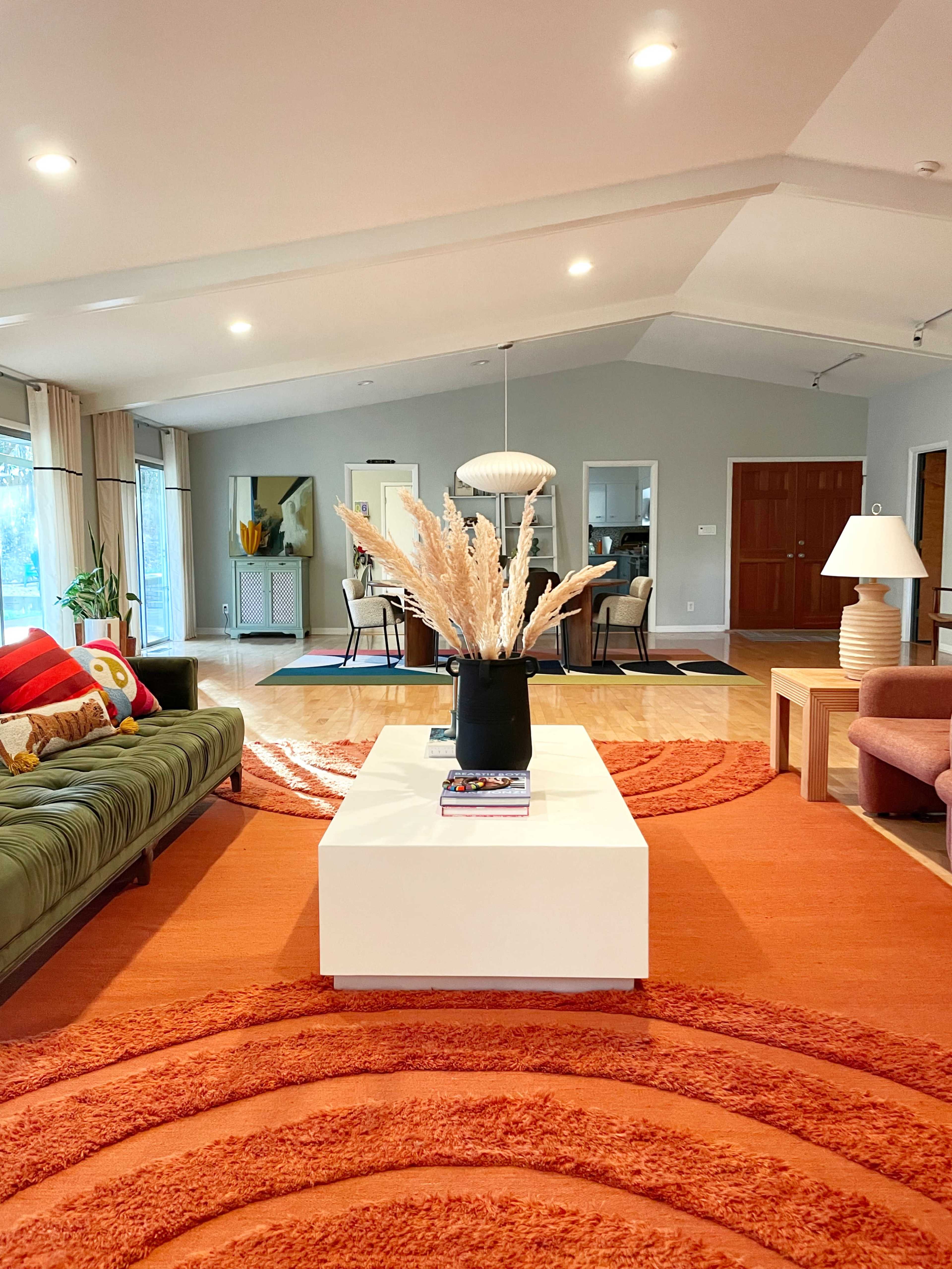 A spacious living room with a green sofa, an orange circular rug, a white coffee table topped with decorative items, and large windows allowing natural light.