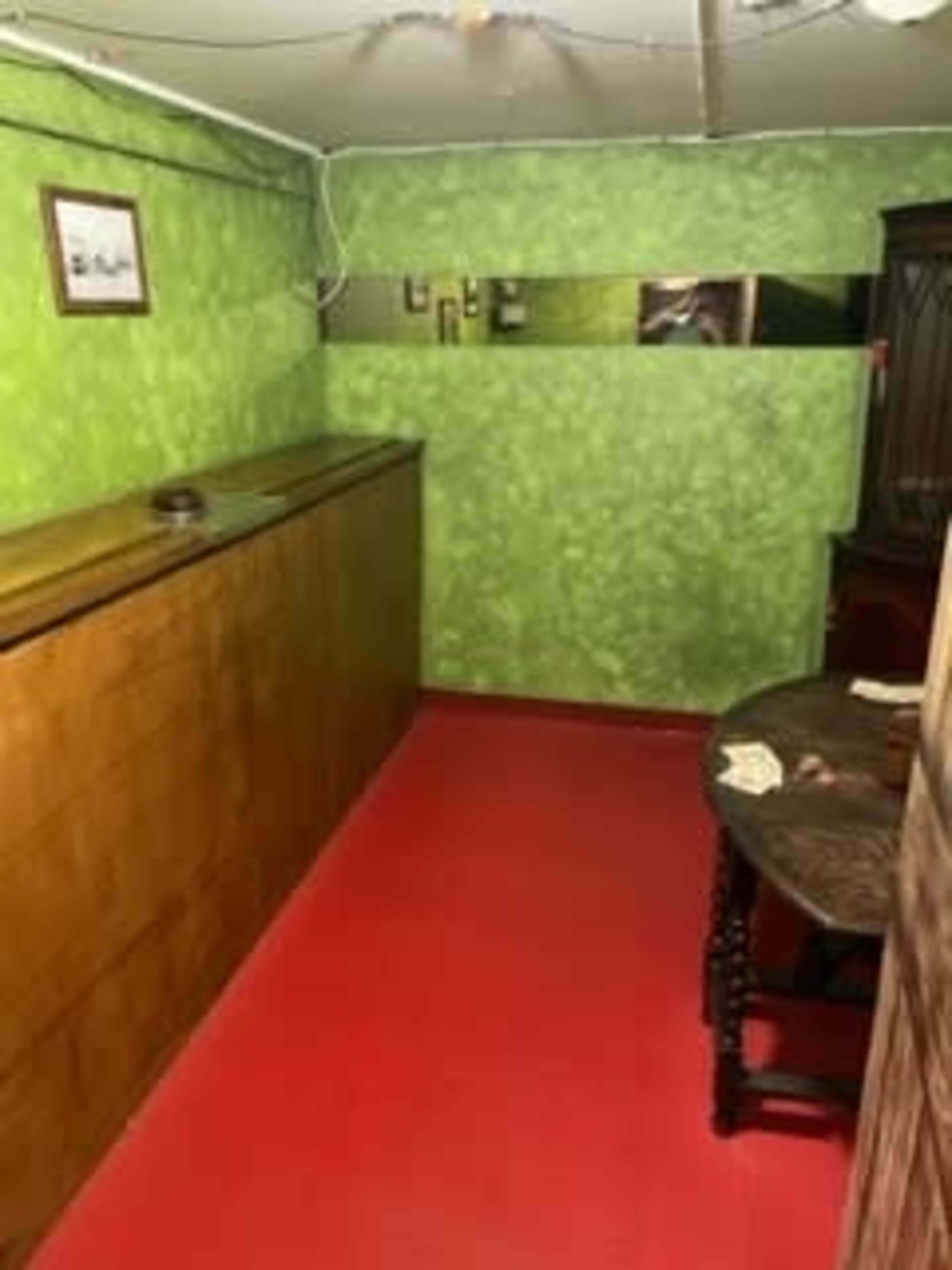The image shows a small room with green walls, a red floor, a wooden table, and a long wooden cabinet against one wall.