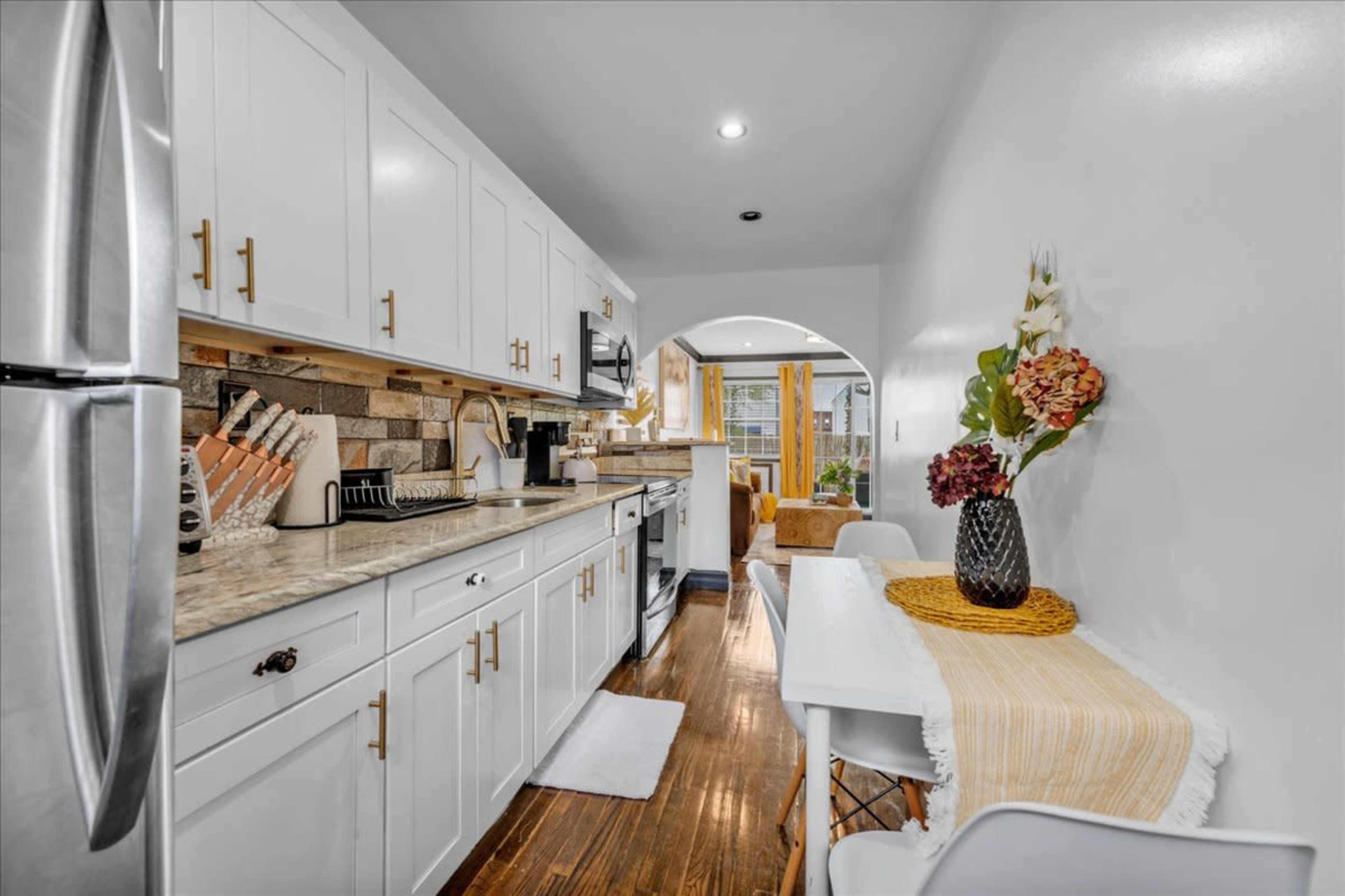A narrow kitchen with white cabinets, a stainless steel refrigerator, and a small dining area near a window.