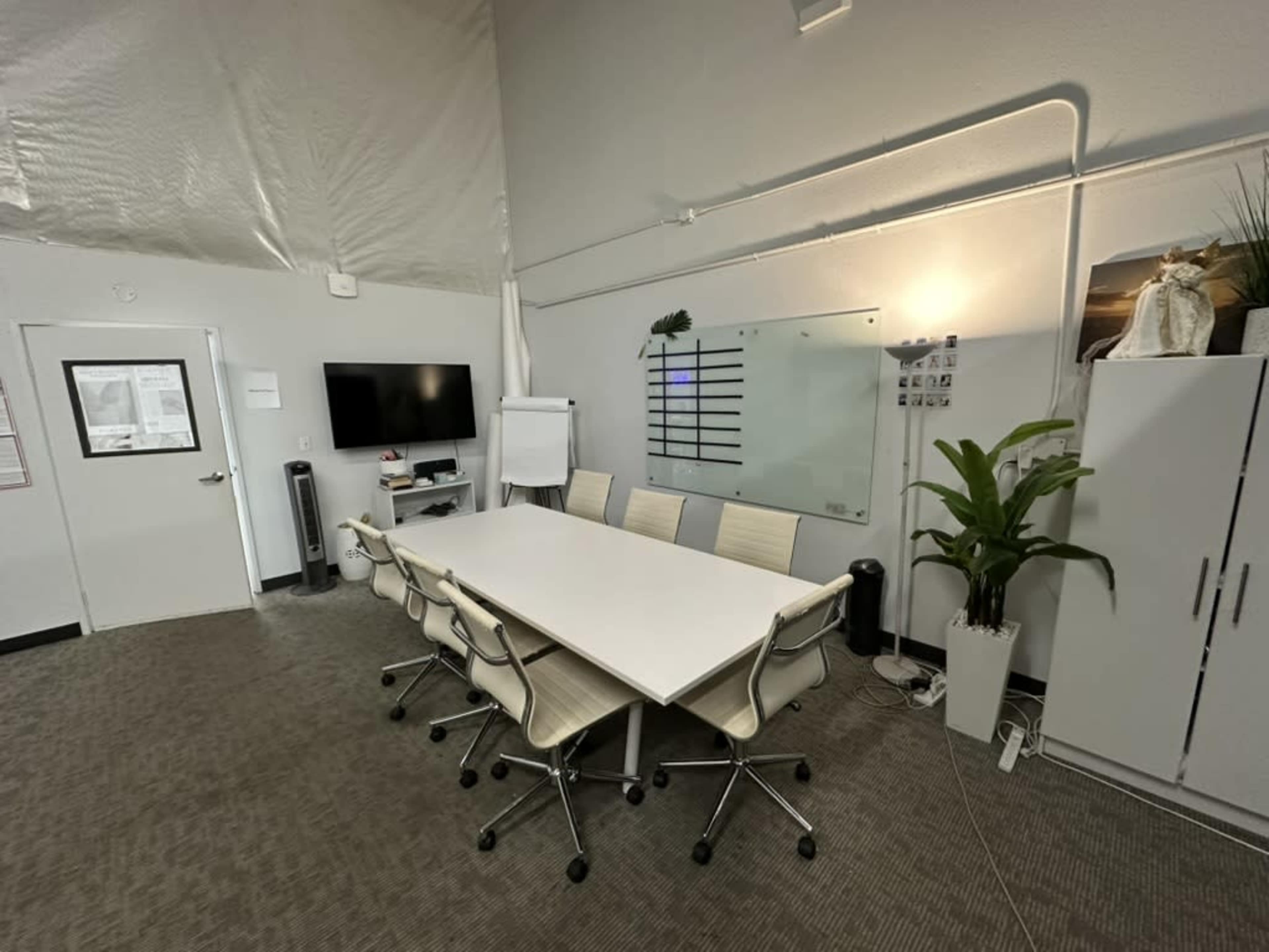 The image shows a modern conference room with a large white table surrounded by six office chairs, a TV mounted on the wall, and a glass whiteboard.