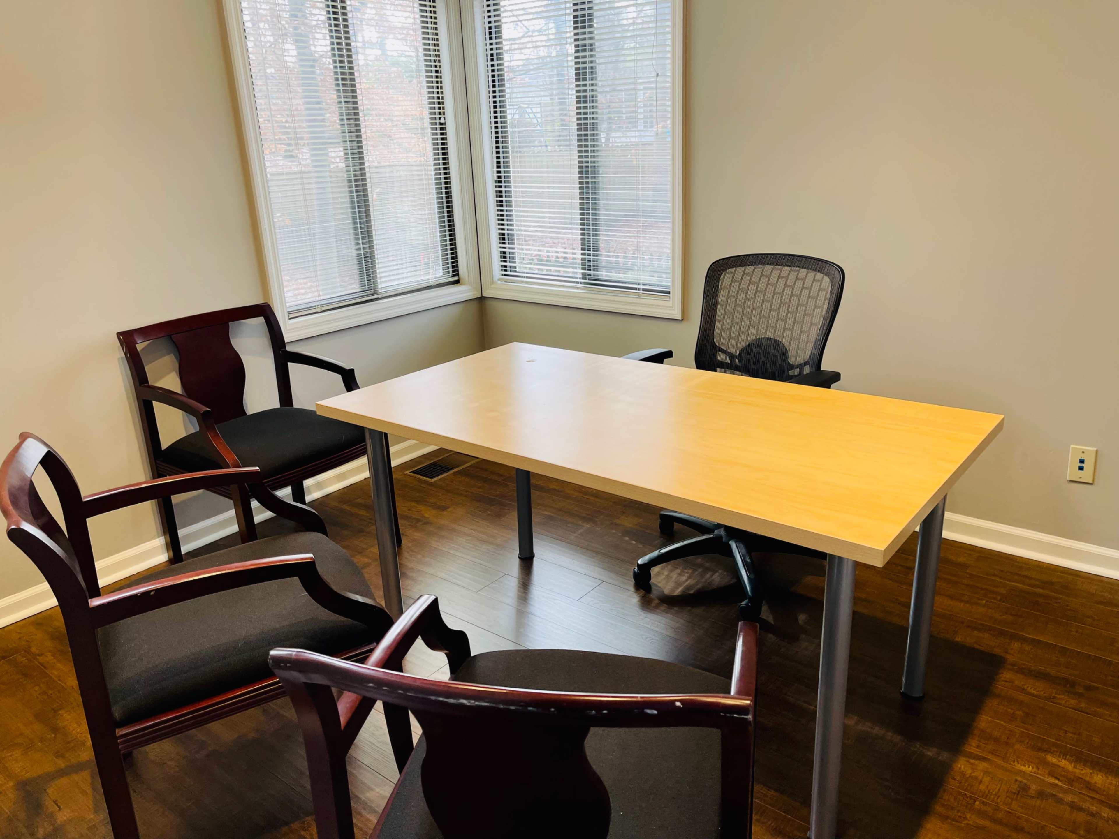 The image shows a meeting room with a wooden table and four chairs, two of which are positioned around the table, and large windows providing natural light.