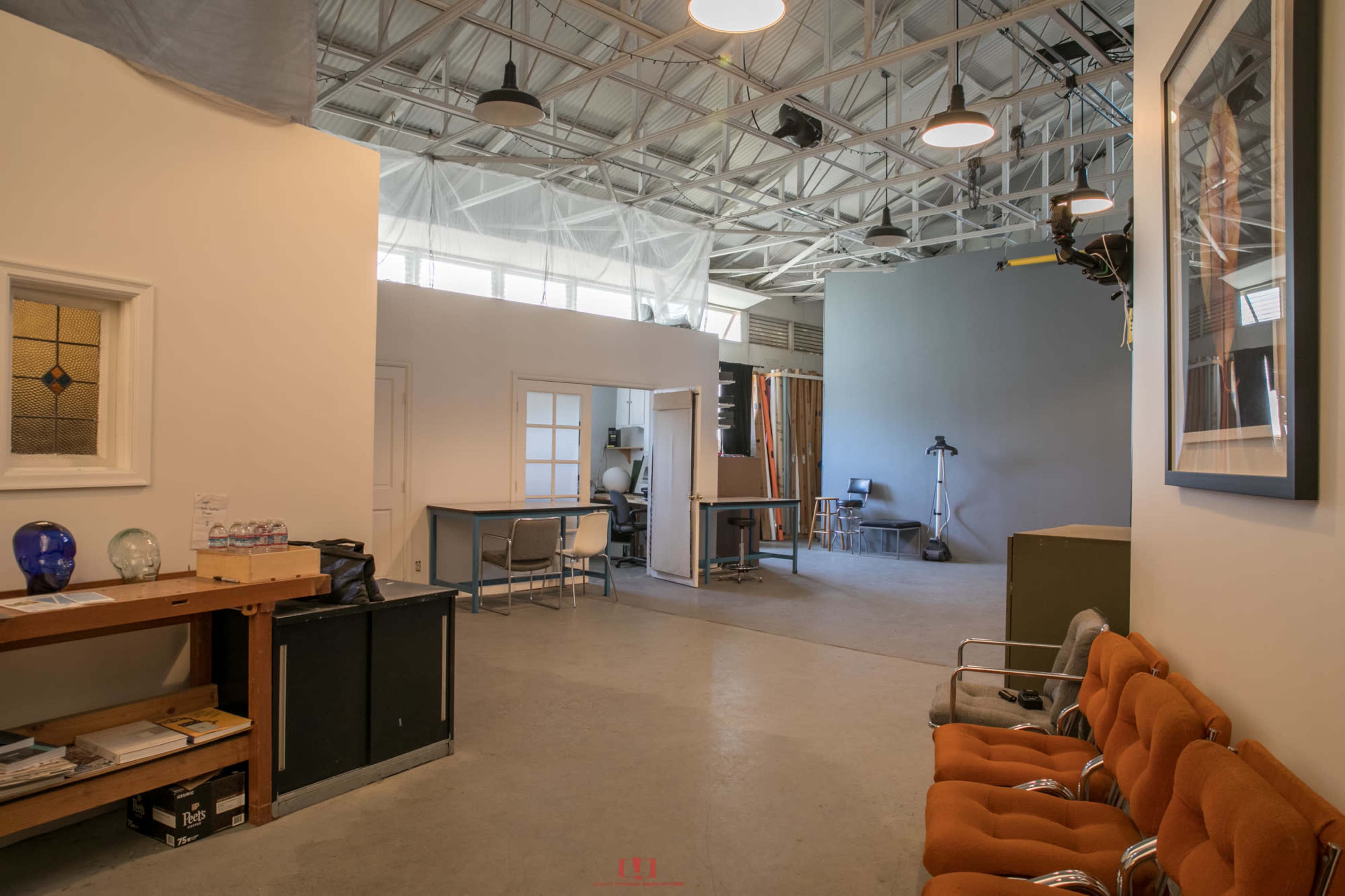 A spacious studio or workshop area with a mix of furniture, including chairs and tables, and large windows allowing natural light to stream in.