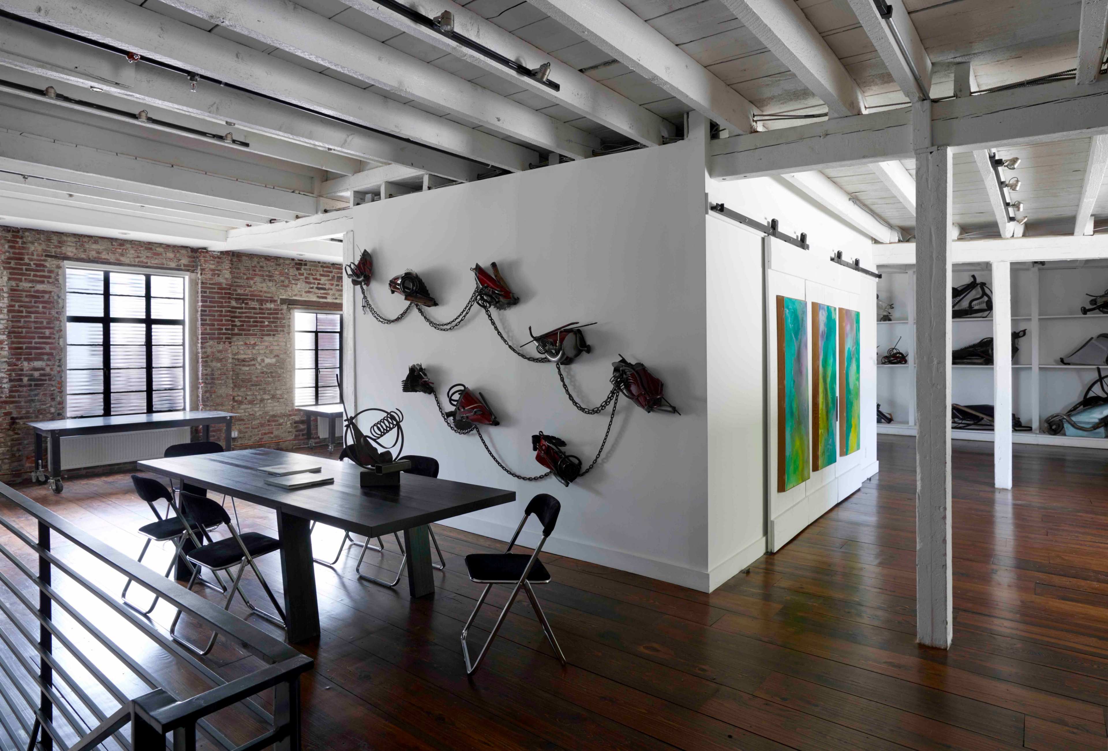 The image shows a modern room featuring a long table with chairs, a white wall adorned with chains and tools, and colorful abstract artwork on another wall, all set in a space with exposed brick and wooden beams.