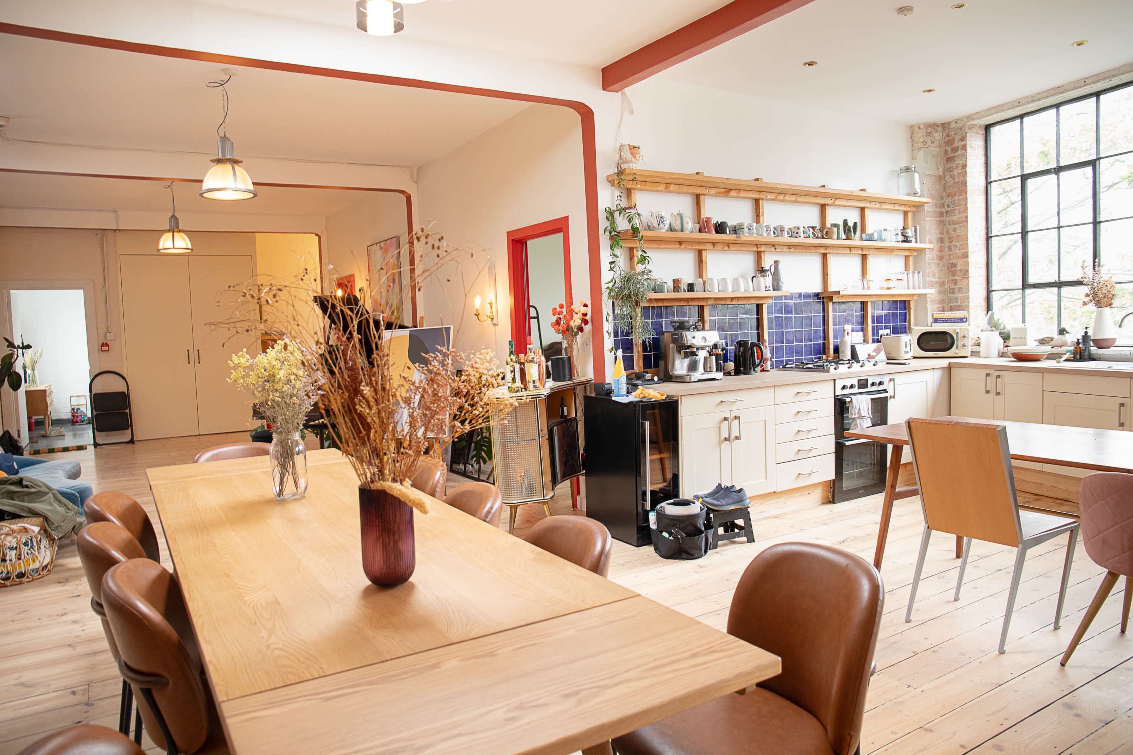 The image shows a spacious, modern kitchen-dining area featuring a long wooden table, stylish chairs, and various kitchen appliances on open shelves.