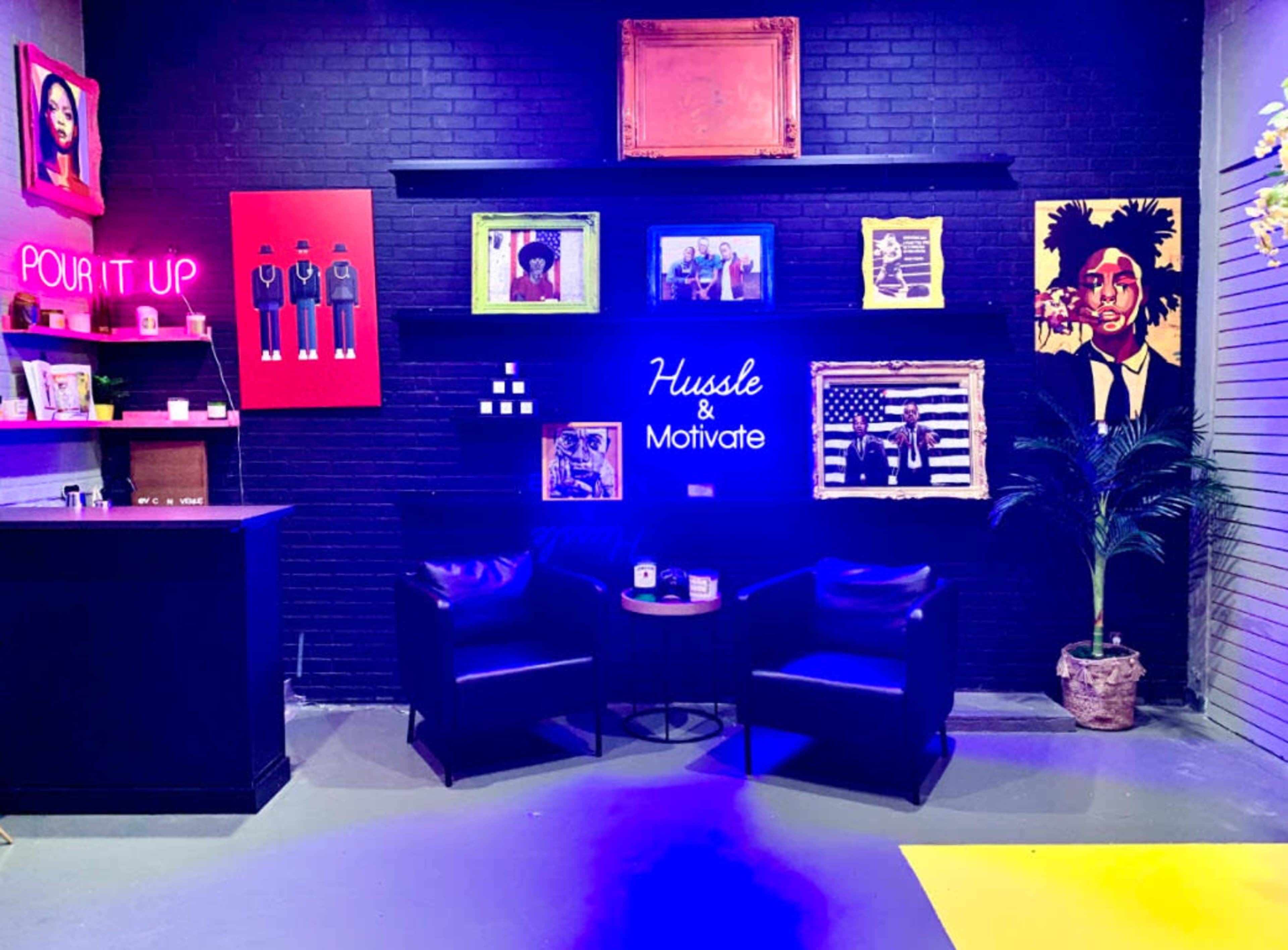 A stylish lounge area featuring black walls adorned with vibrant art, a neon sign reading "Hussle & Motivate," and two black chairs beside a small table.