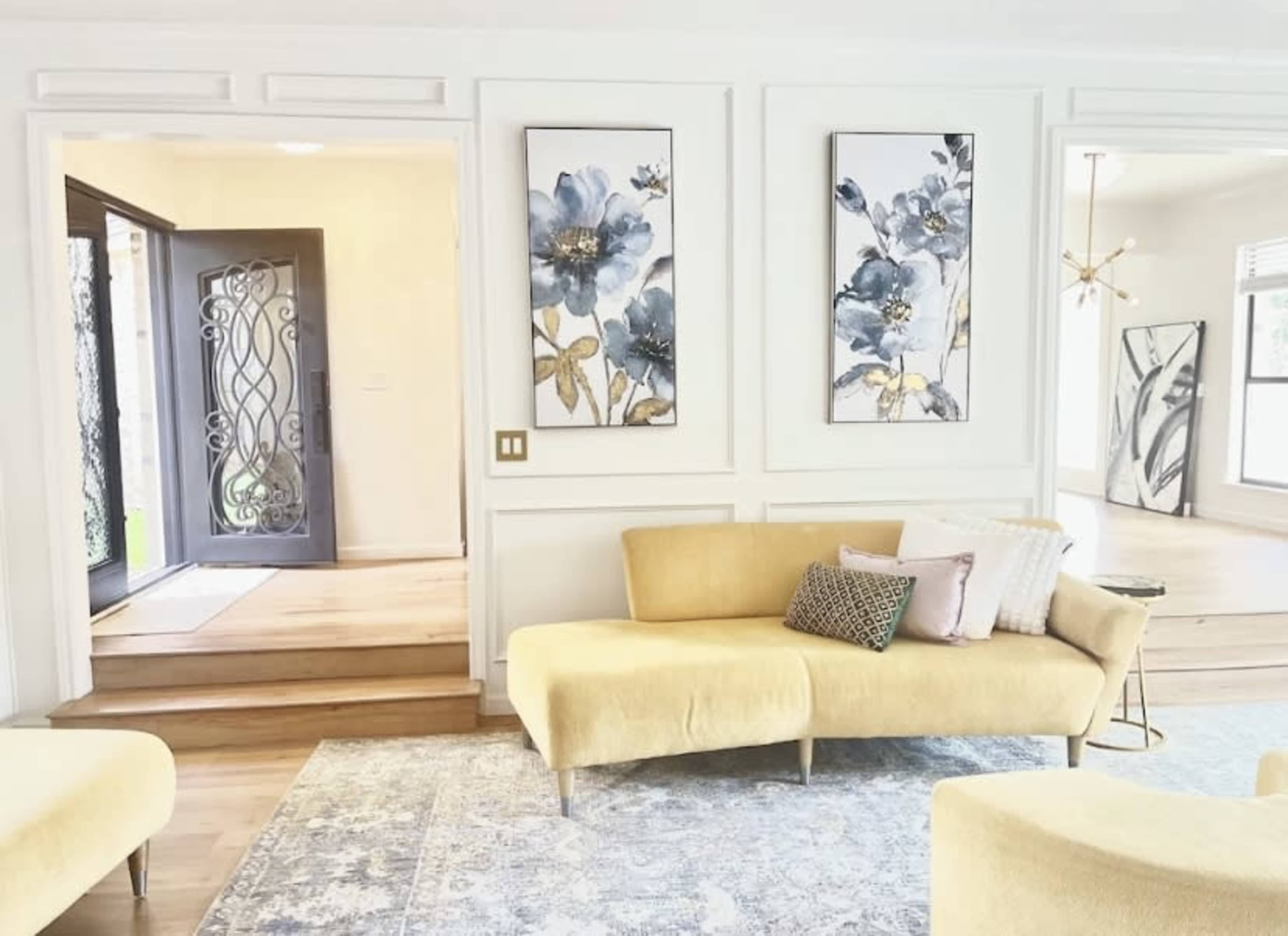 The image shows a light-colored living room with a yellow sofa, decorative wall art featuring floral designs, and a front door opening to another area.