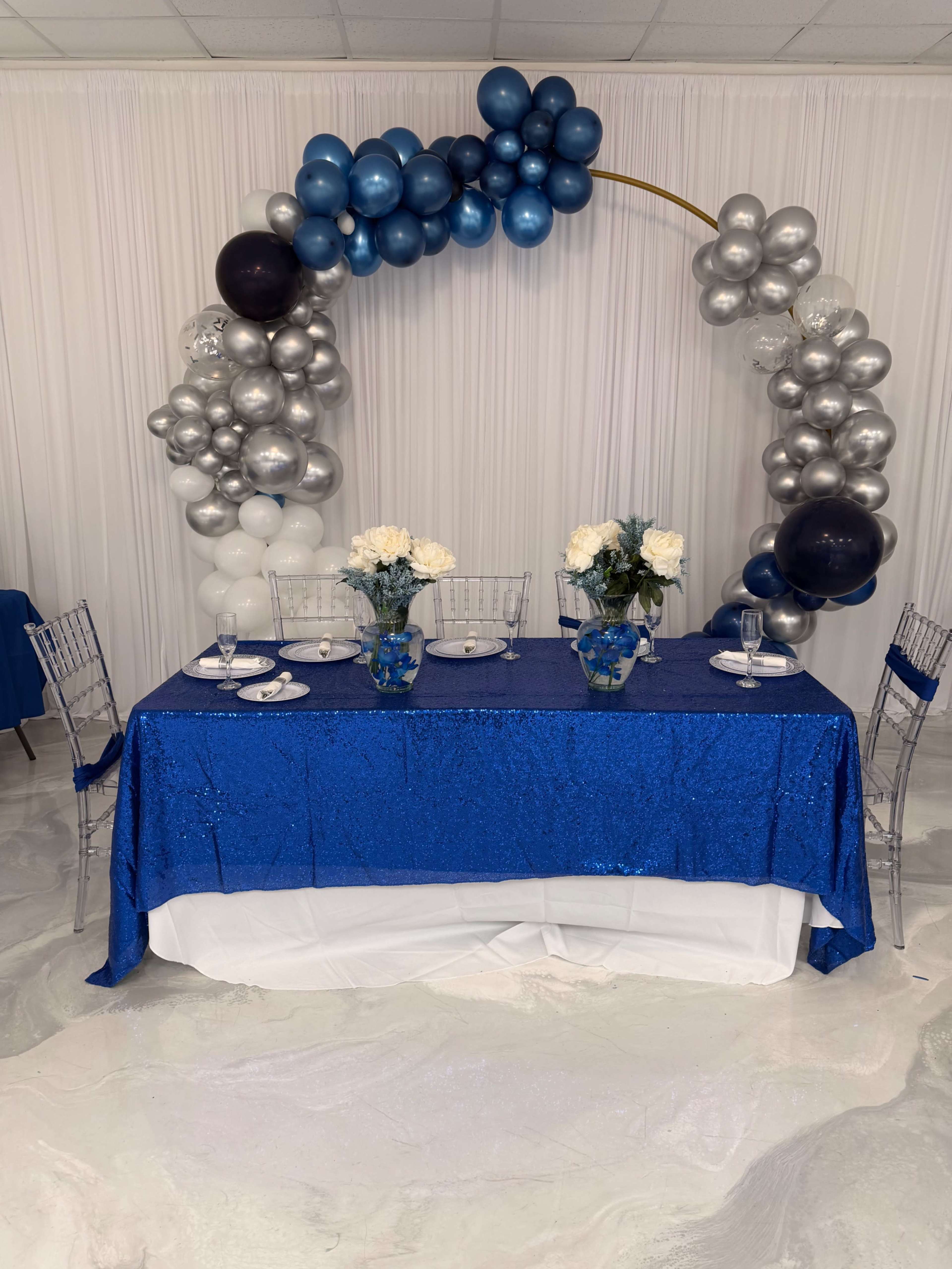 The image shows a decorated table with a blue sequin tablecloth, two vases with white flowers, and a balloon arch made of blue, silver, and black balloons in the background.