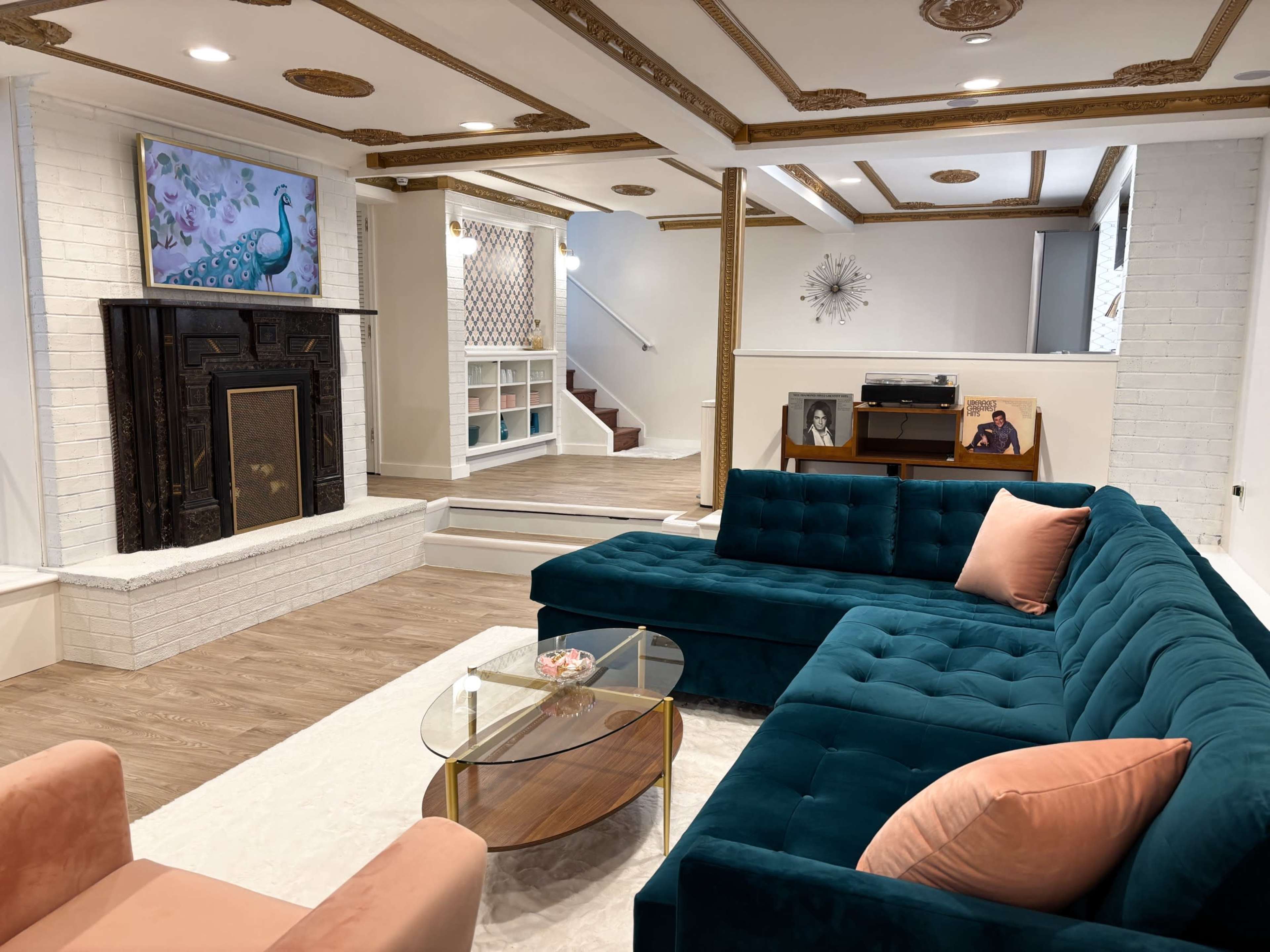 The image shows a spacious living room featuring a teal sectional sofa, a glass coffee table, a fireplace with a decorative mantle, and a textured wall with a peacock artwork.