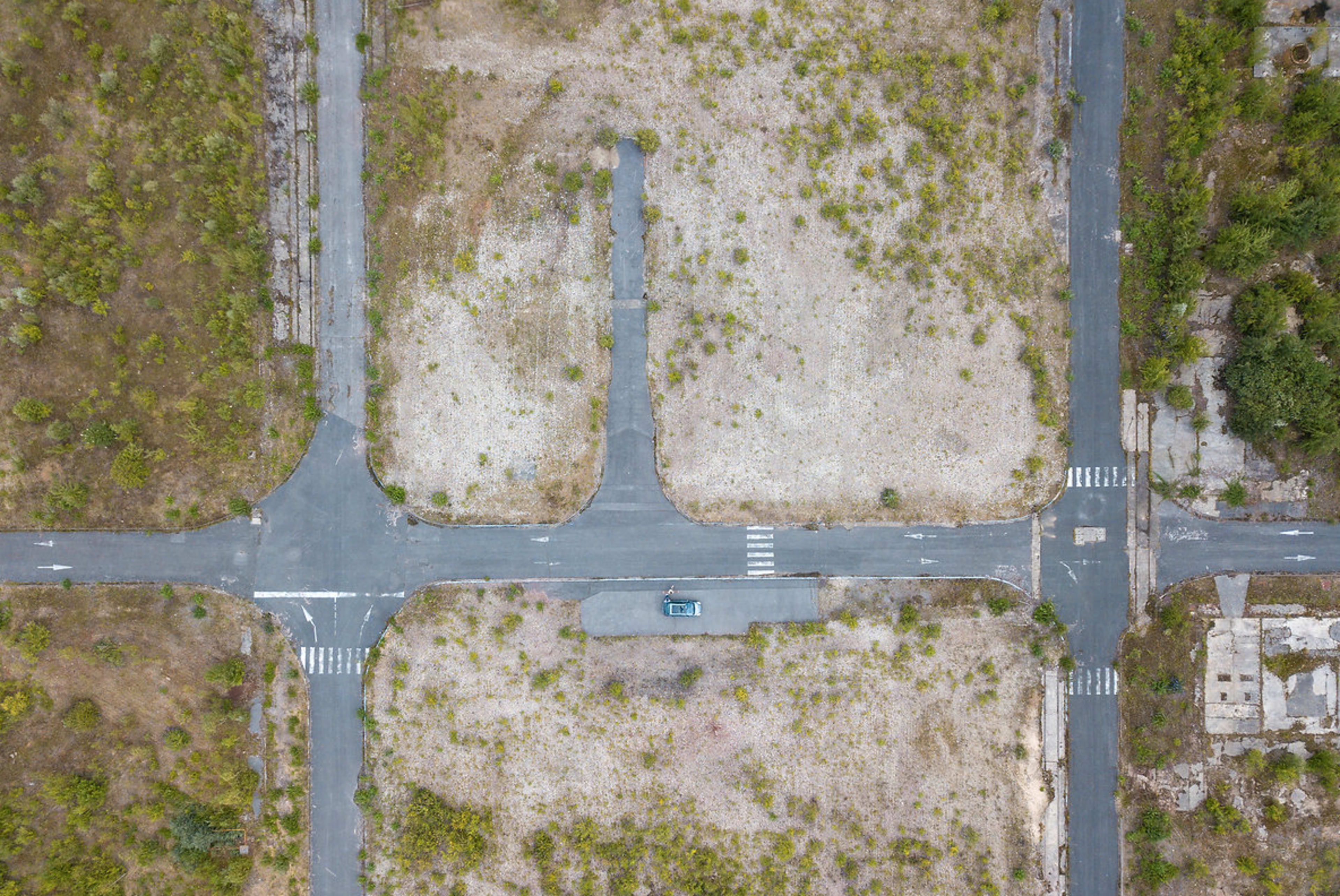 An aerial view shows a vacant intersection surrounded by overgrown vegetation and sparse asphalt roads.