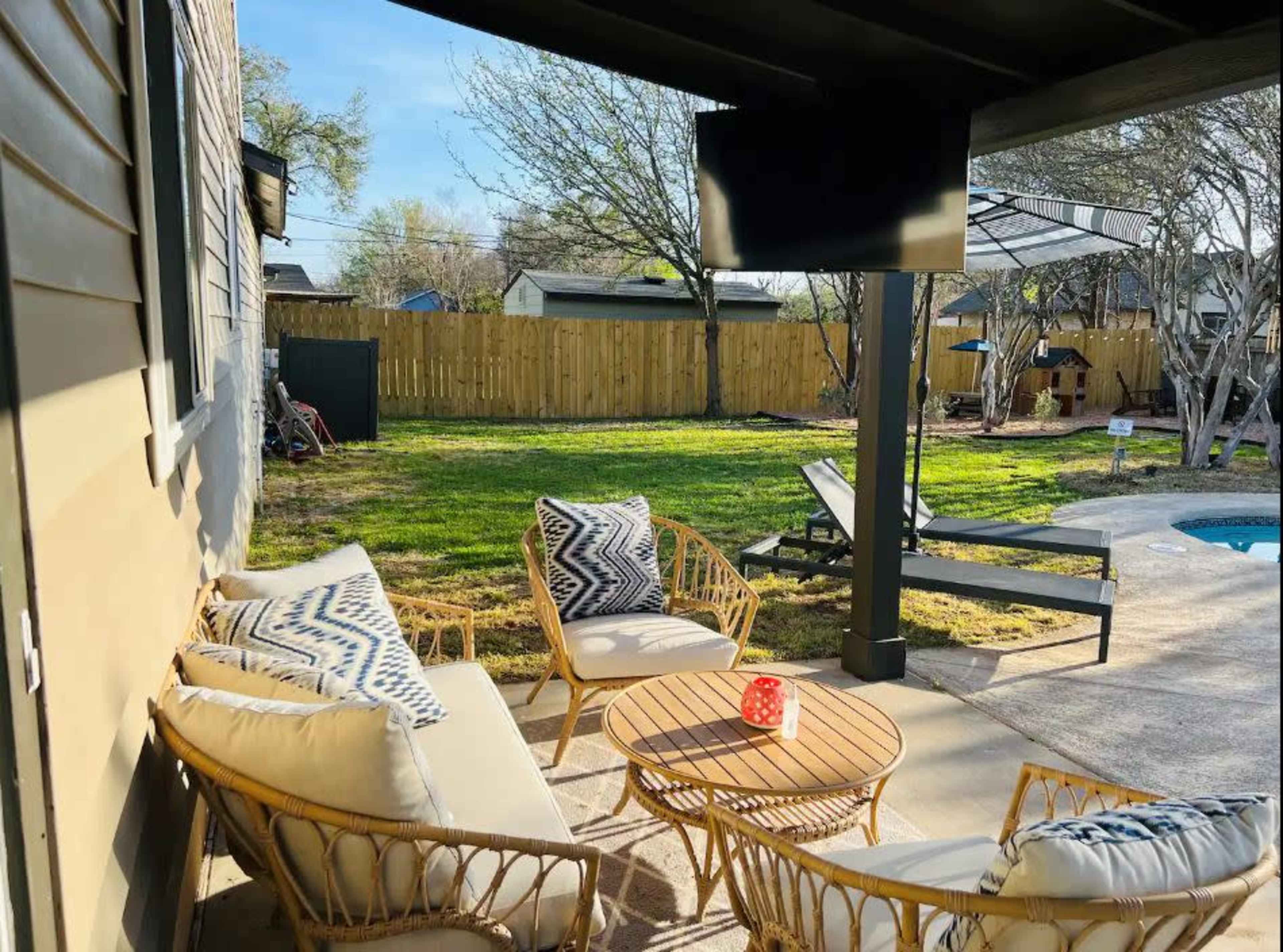 A patio area features a seating arrangement with wicker furniture, a small table, and a mounted television, overlooking a green backyard with trees and a pool.