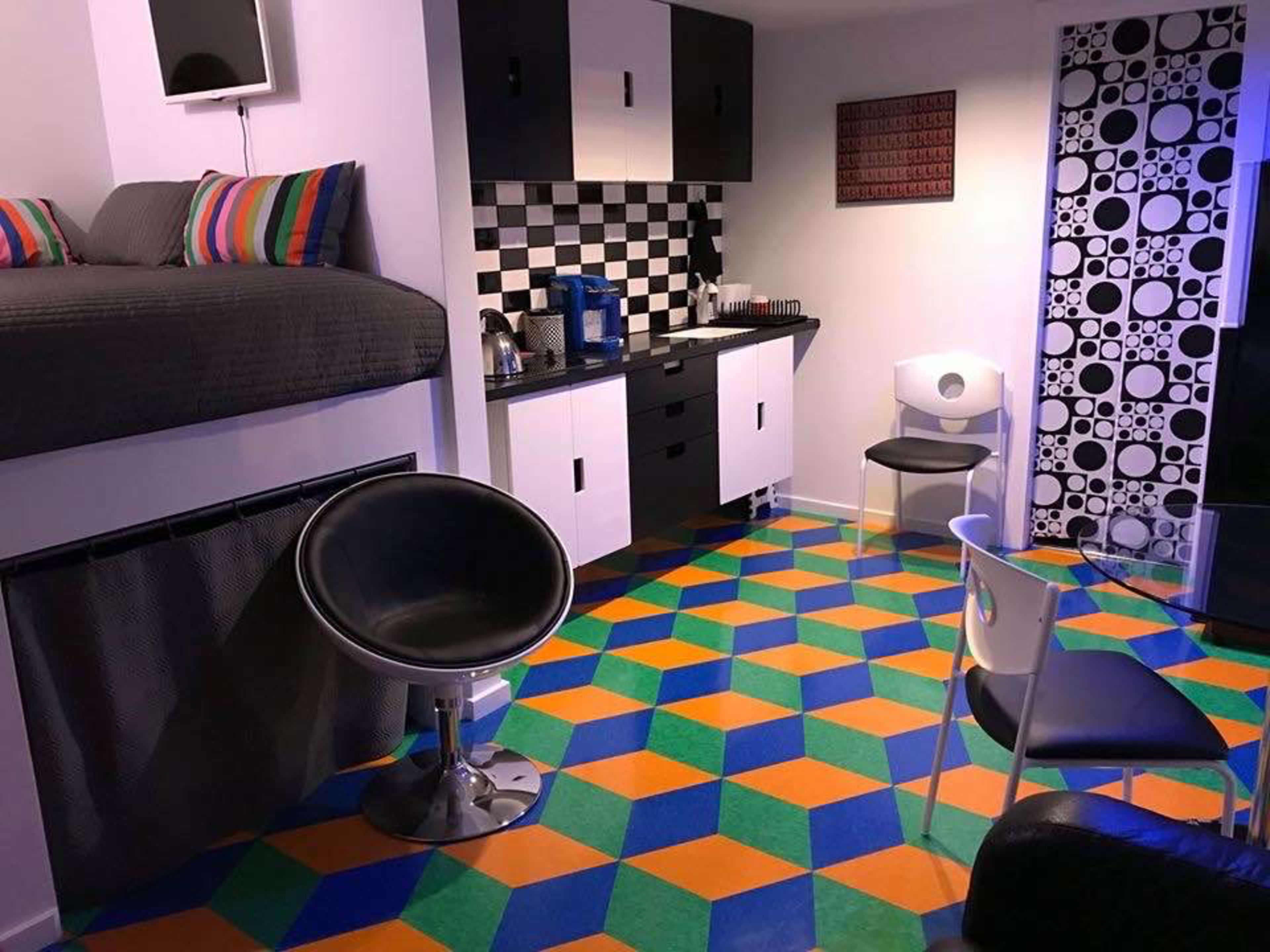 A small, modern room featuring a lofted bed, a checkered kitchen area, and a colorful geometric-patterned floor.
