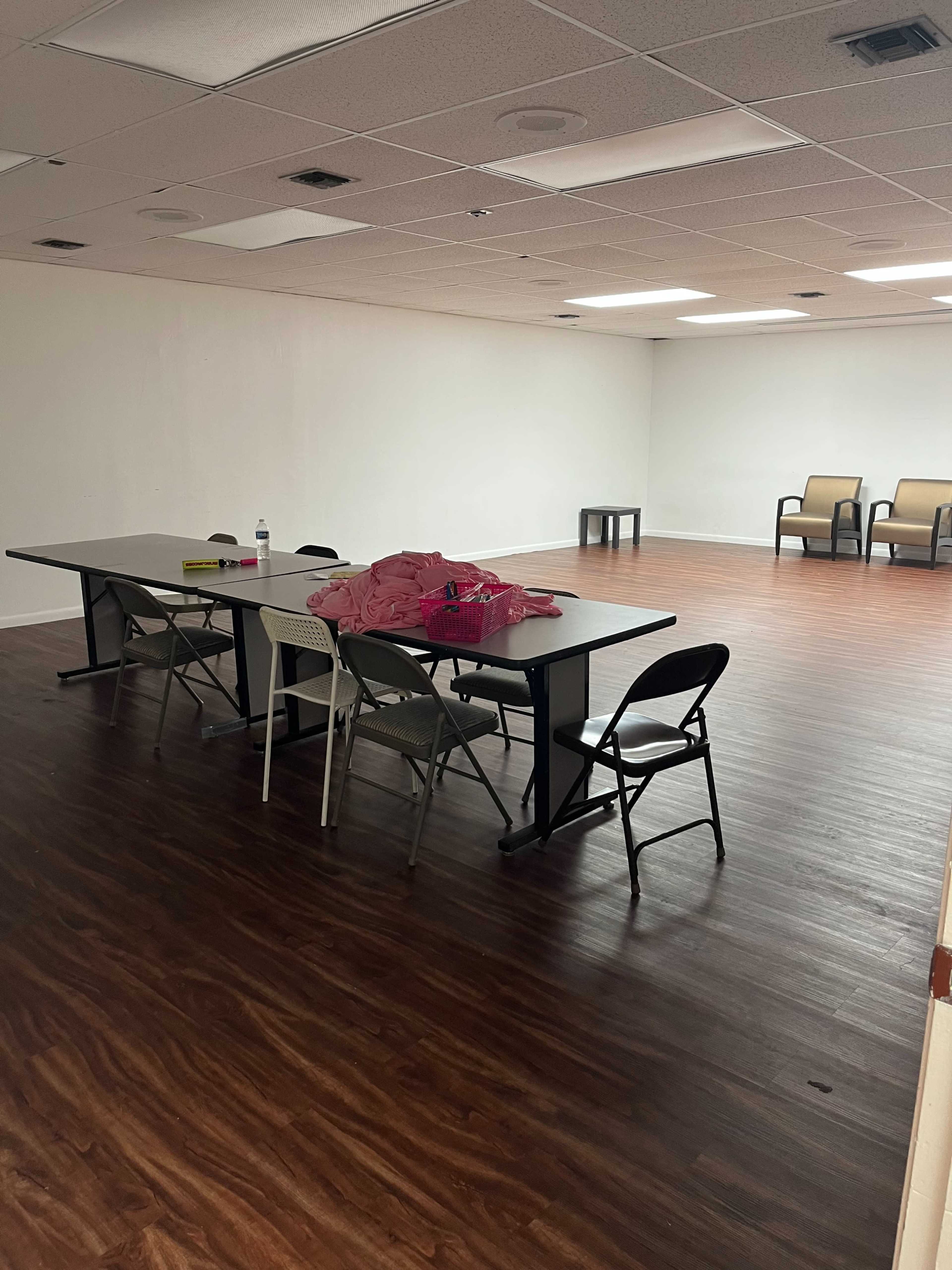 A sparsely furnished room with a long table surrounded by chairs and two empty seating areas in the background.