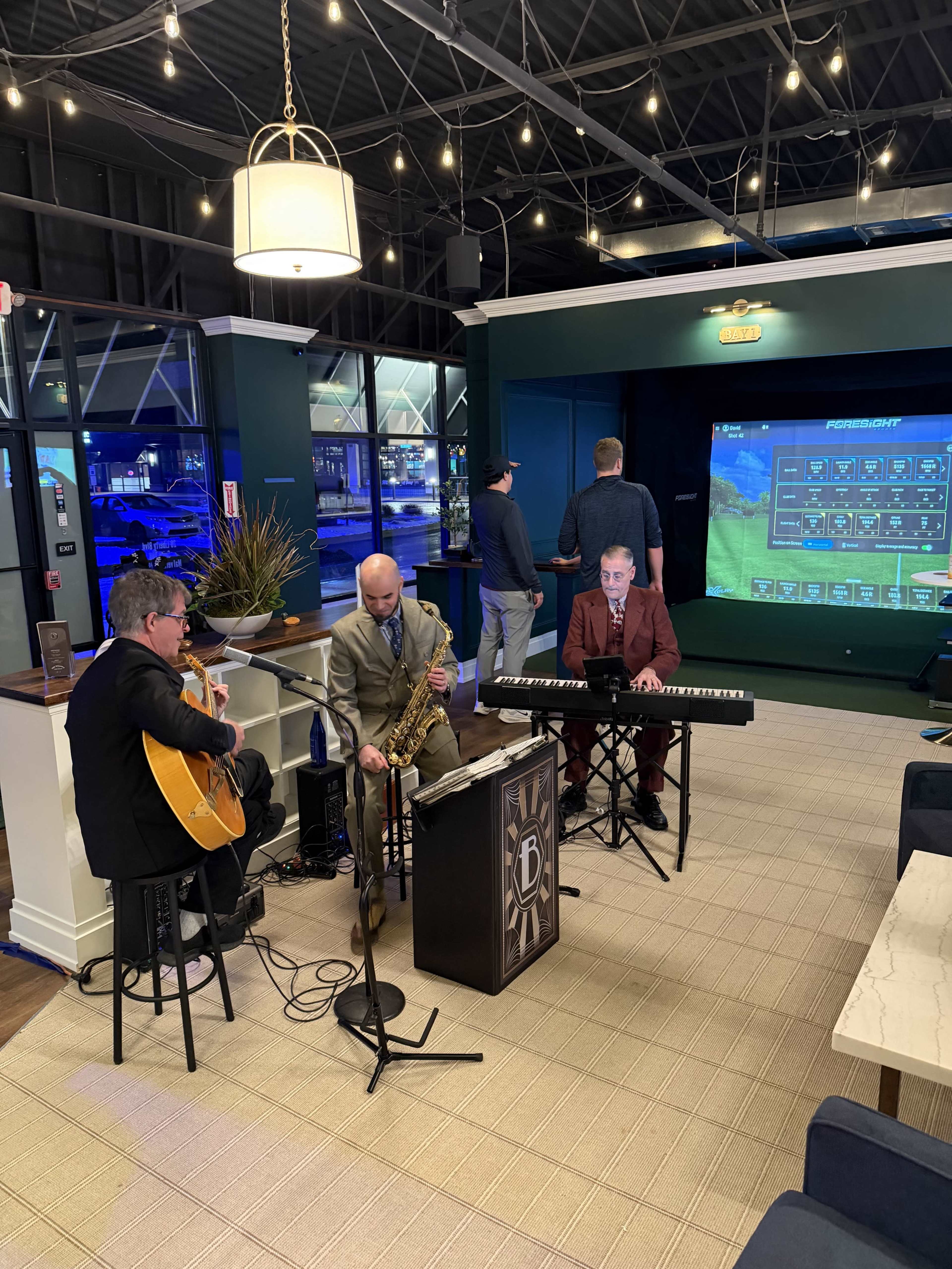 A trio of musicians performs in a modern lounge setting, with a saxophonist, guitarist, and keyboardist in front of a large screen displaying graphics.