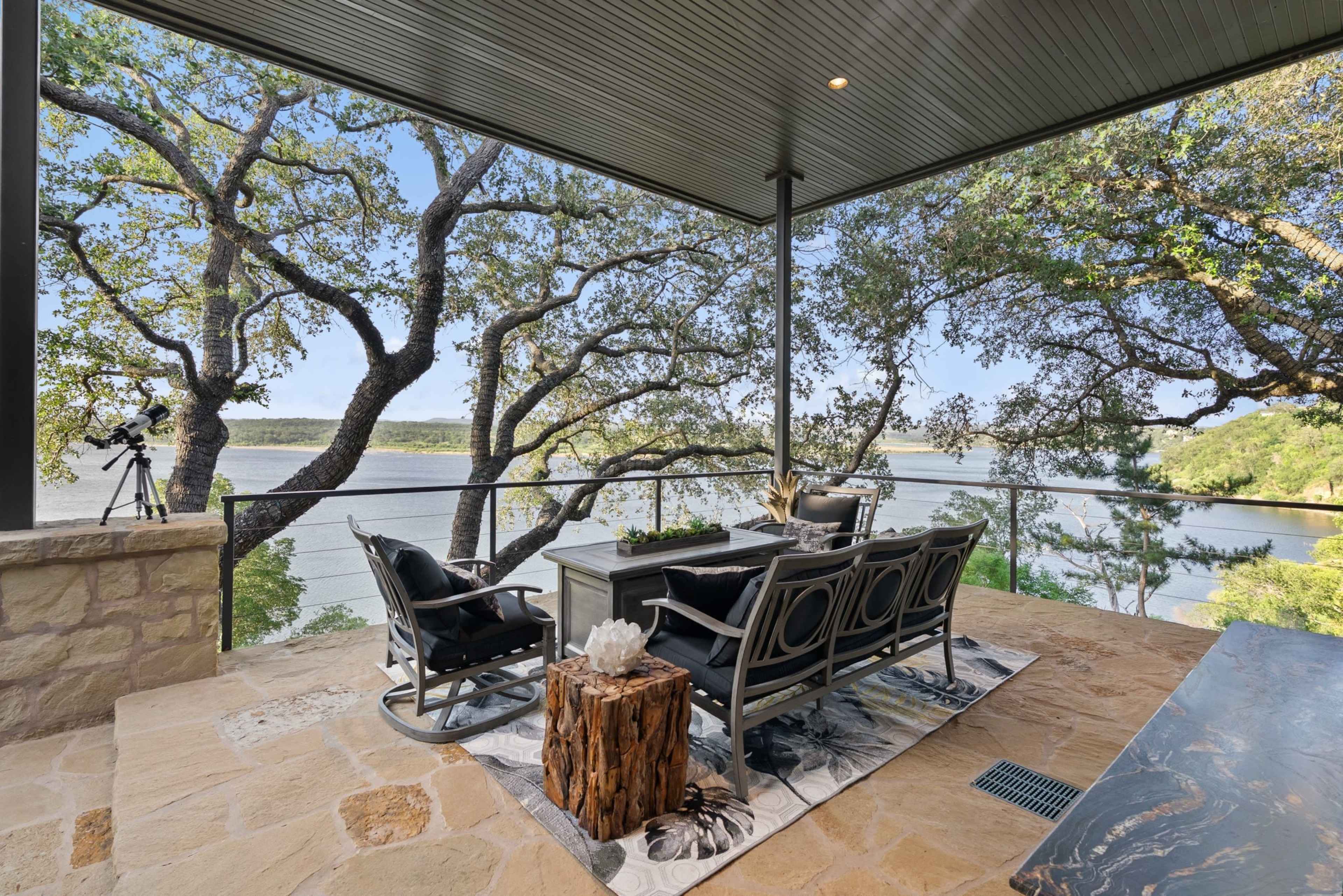 A patio overlooking a lake features a seating area with a table, surrounded by trees.