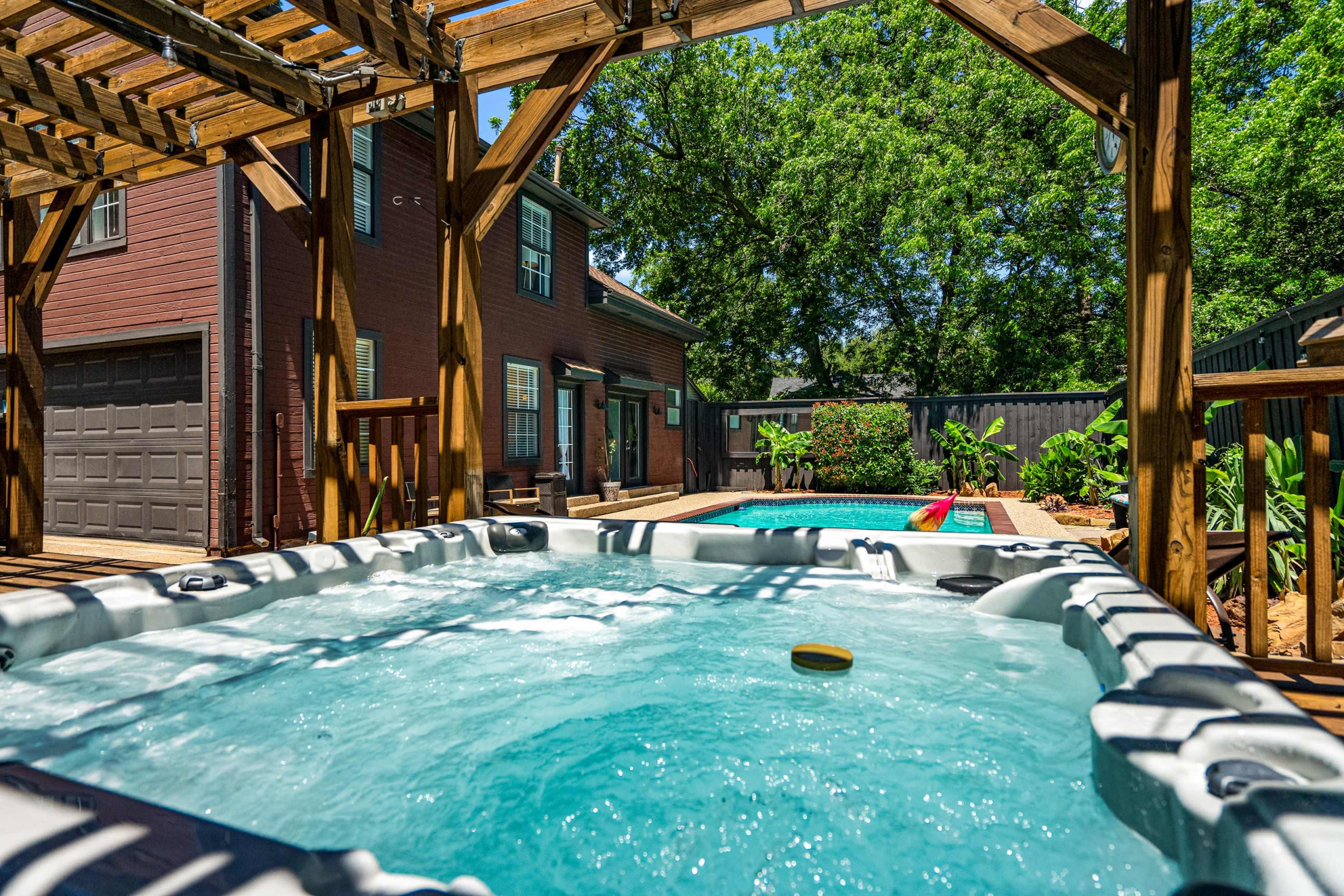 A hot tub is situated near a swimming pool in a backyard surrounded by trees and a two-story house.