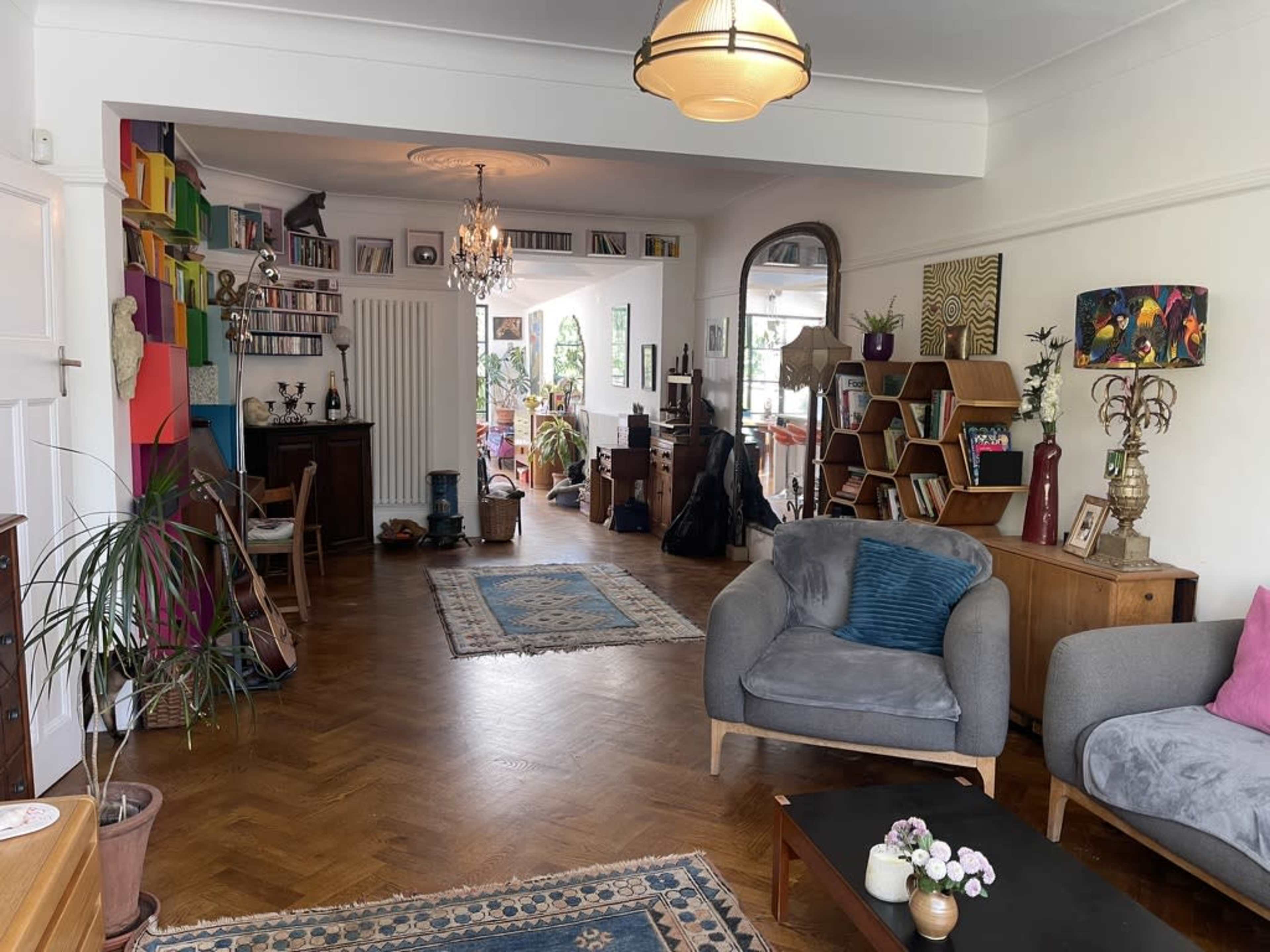 The image shows a spacious living room with wooden flooring, bookshelves, colorful wall art, and various seating arrangements including a gray sofa and a coffee table.