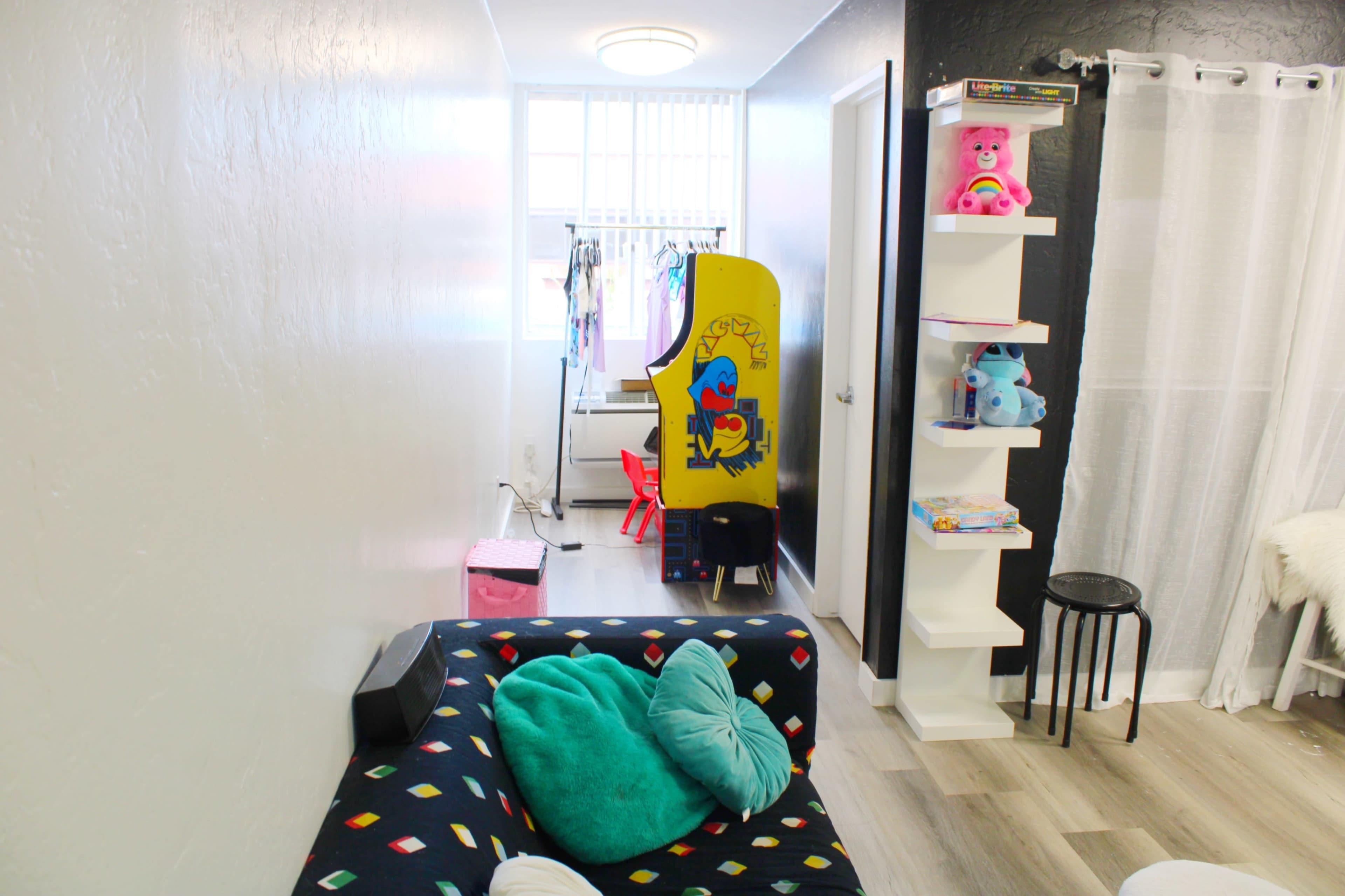 The image shows a narrow hallway with a colorful couch, a vintage arcade game cabinet, a clothing rack, and a shelving unit containing toys and books.