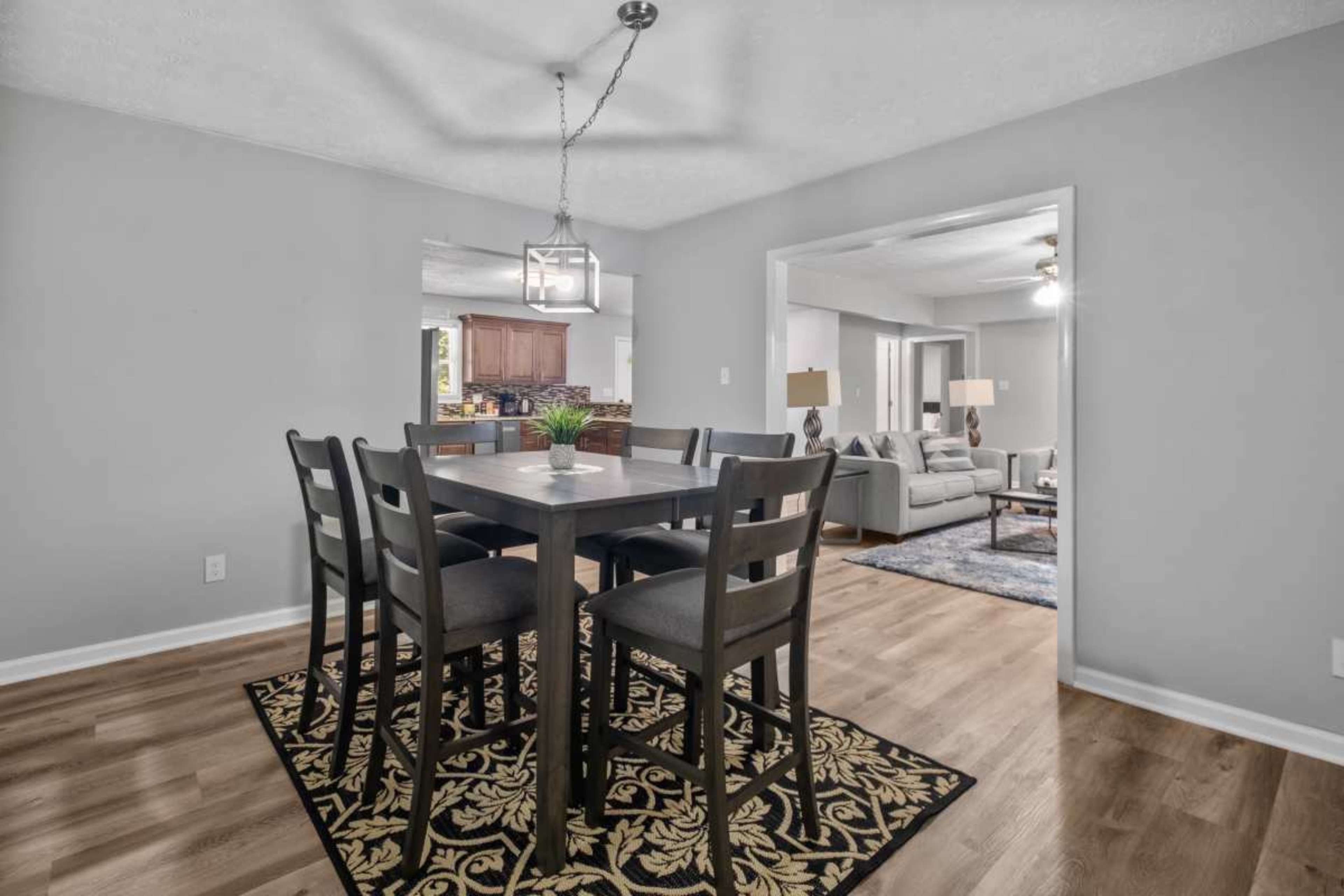 A dining area features a square table surrounded by tall chairs, with a rug underneath and an open view into a living space.