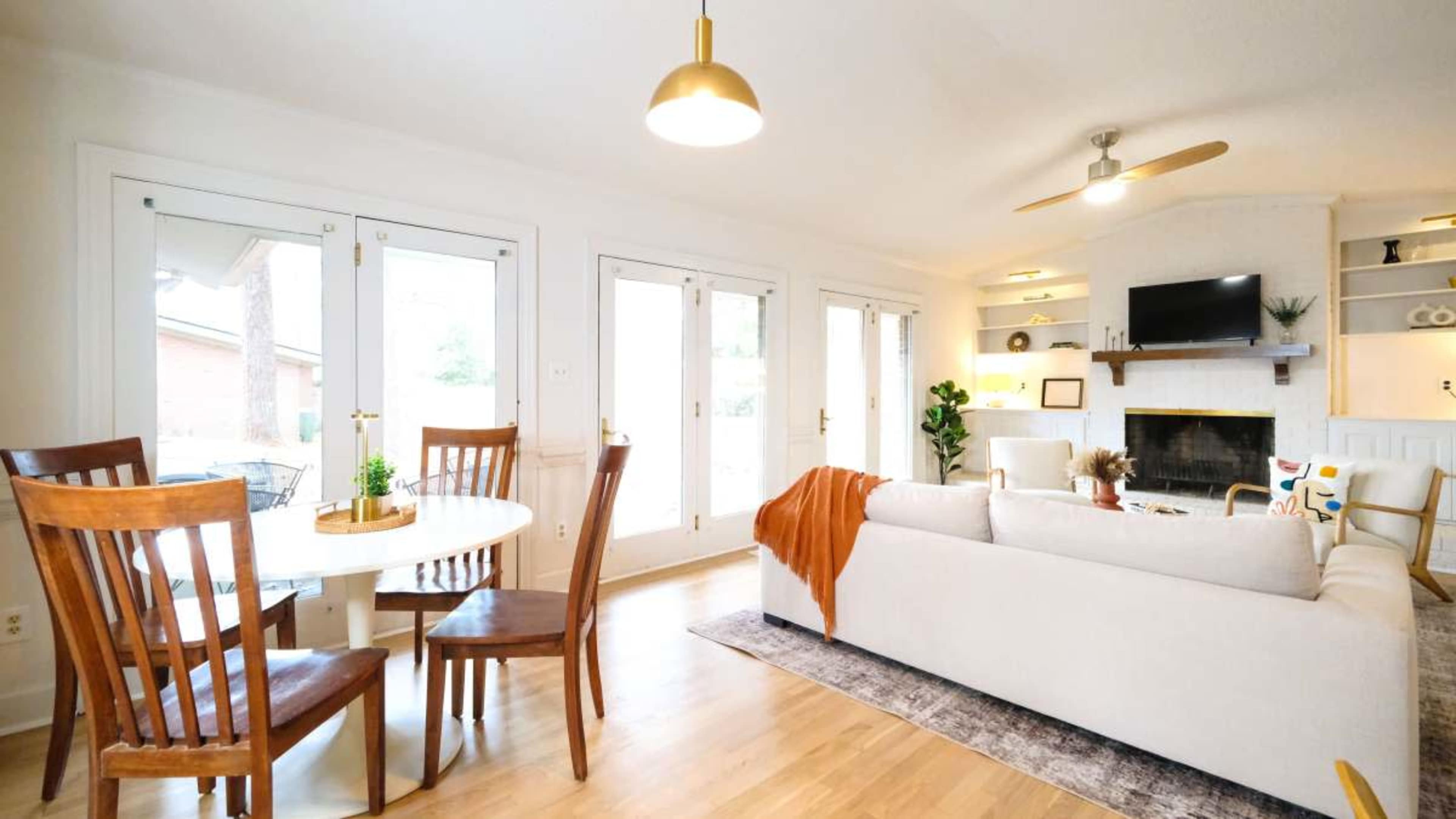 The image shows a modern living room with a light-colored couch, a circular dining table with wooden chairs, and large windows leading to an outdoor space.