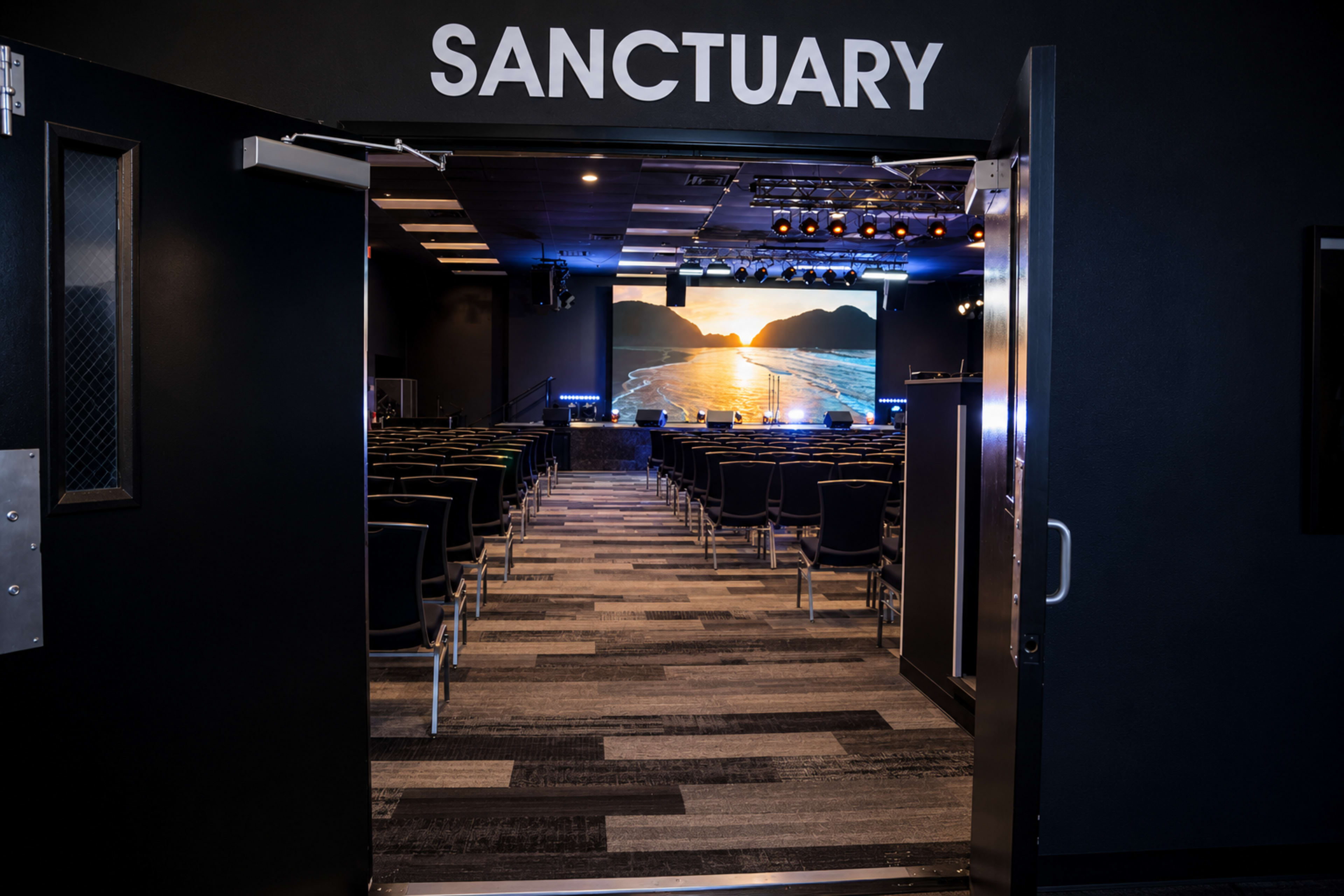 The image shows an entrance to a room labeled "SANCTUARY," leading into a spacious area filled with rows of chairs and a stage displaying a scenic backdrop of a sunset over mountains.