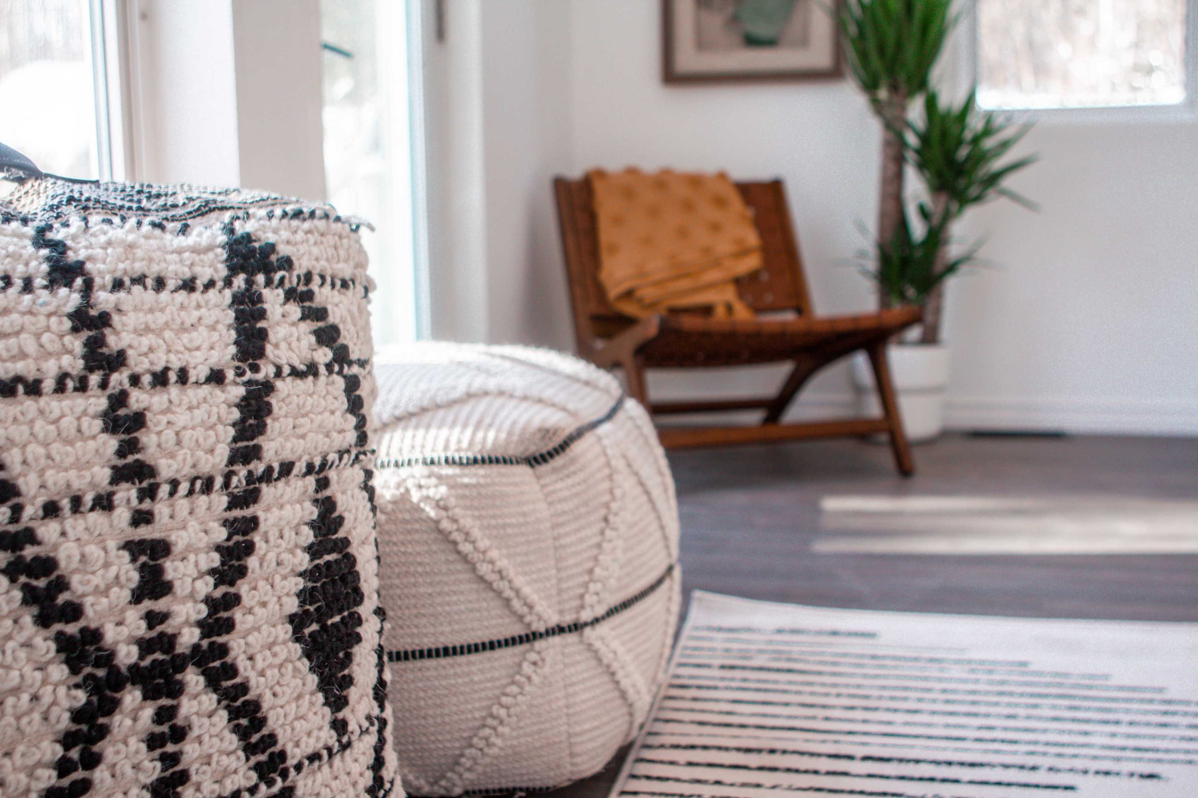 A cozy living space featuring a patterned pouf in the foreground, a wooden chair with a blanket, and a potted plant in the background.