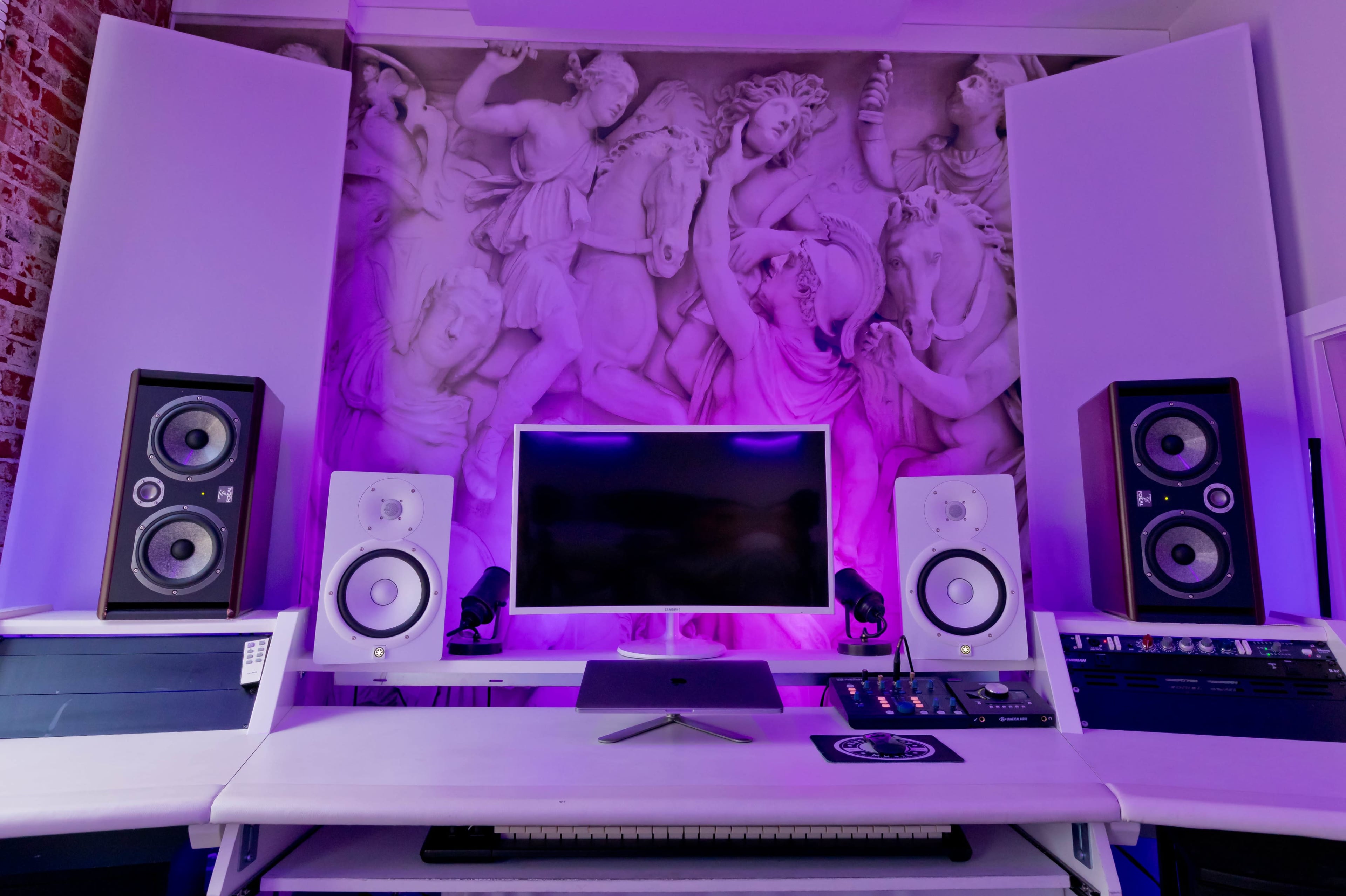 A music production setup features a central monitor flanked by studio speakers, with a decorative wall mural in the background.