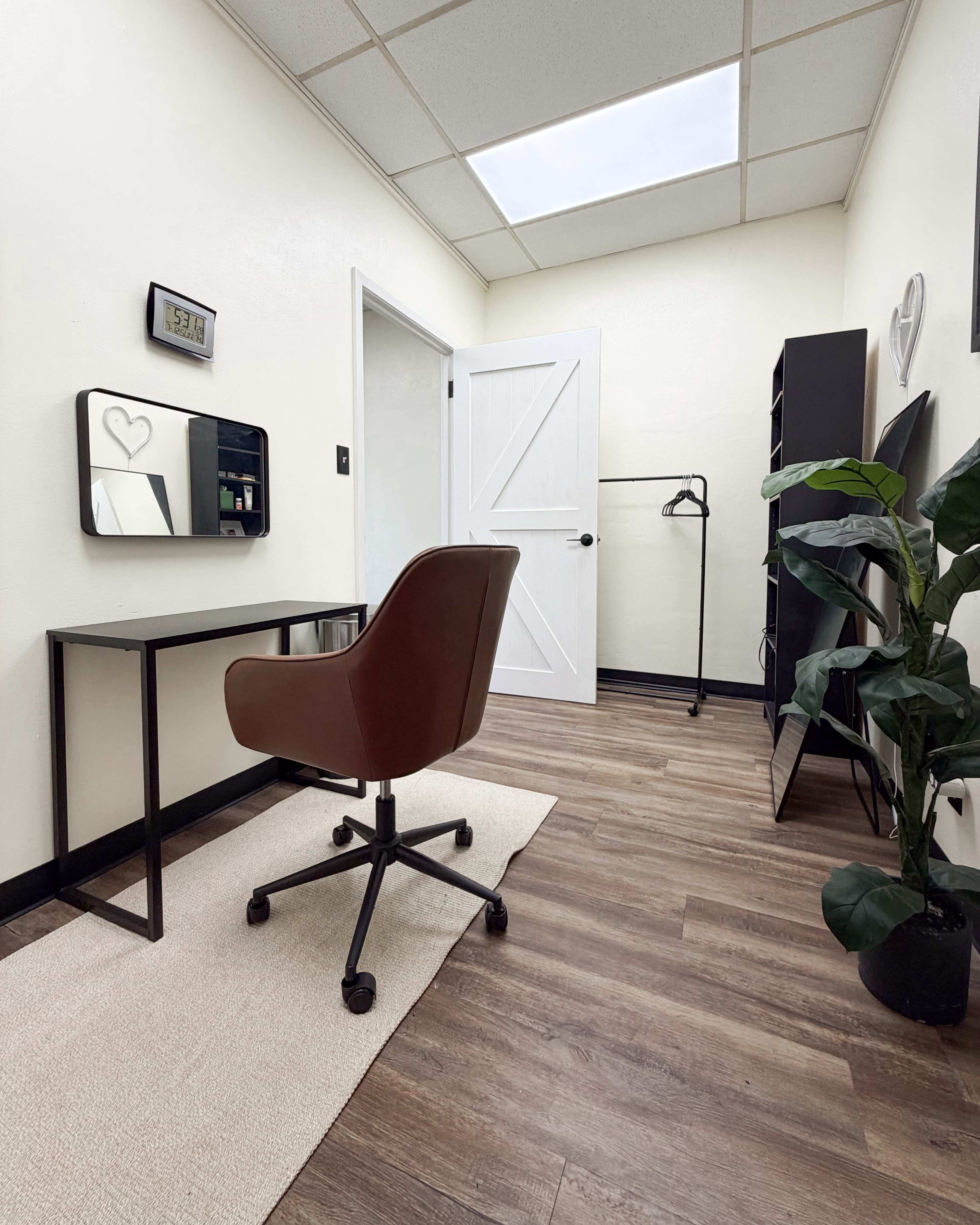 The image shows a small, tidy office space featuring a desk with a chair, a mirror, a clothing rack, and a plant in a corner.