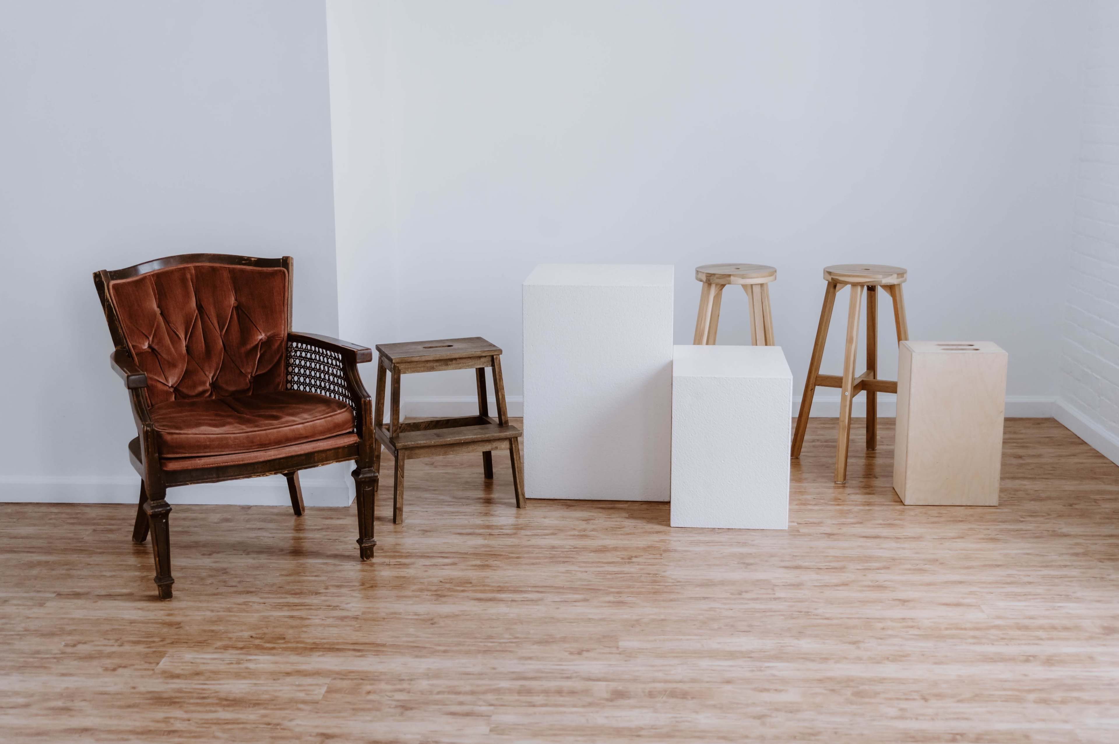 A room with wooden flooring, a brown upholstered armchair, two white blocks, a small wooden step stool, and three wooden stools.