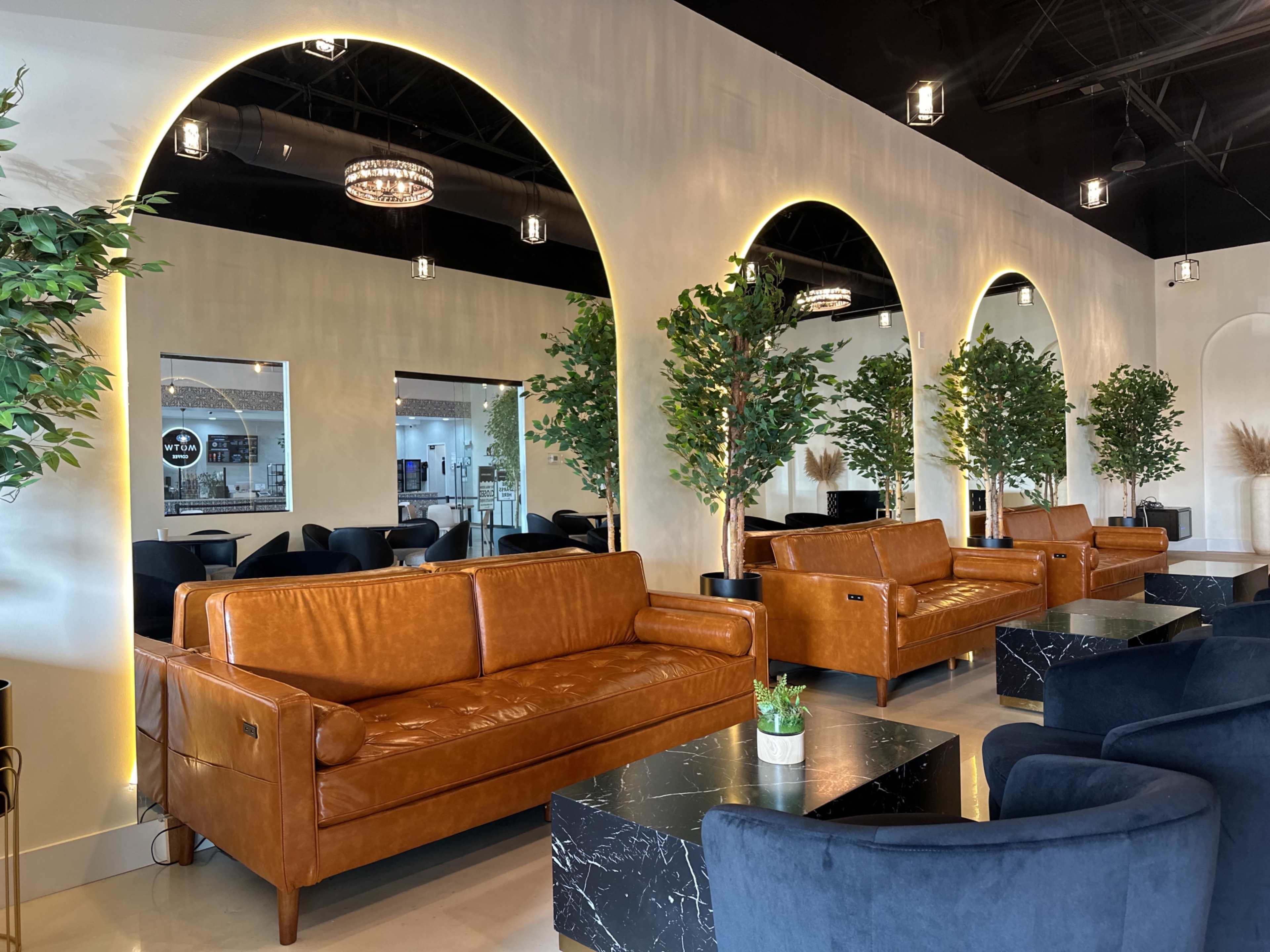 The image features a modern lounge area with brown leather sofas and dark chairs arranged around black marble tables, flanked by greenery and illuminated arches along the walls.