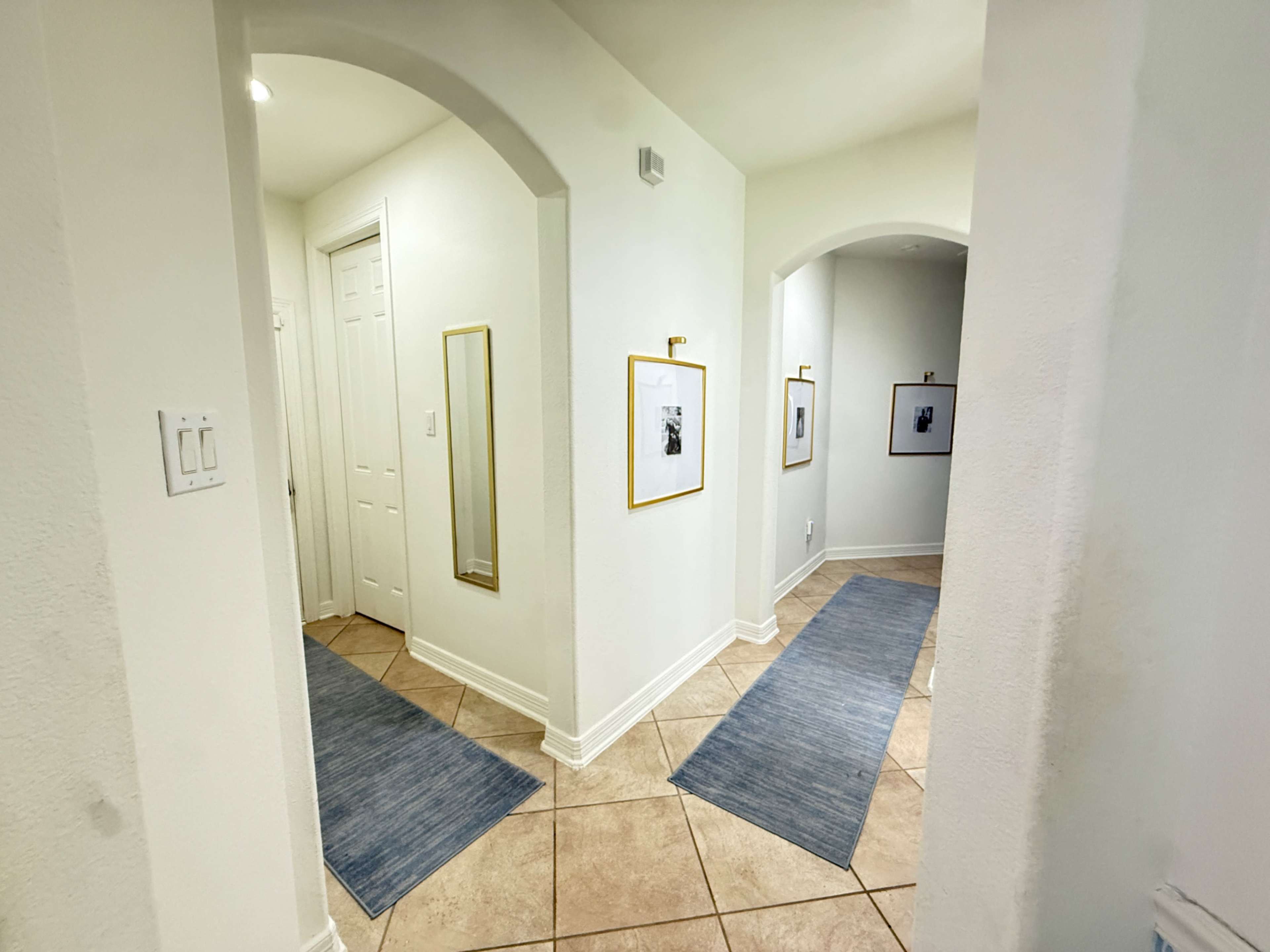 The image shows a well-lit hallway with arched doorways, light-colored walls, and blue runners on the tiled floor, featuring framed pictures on the walls.