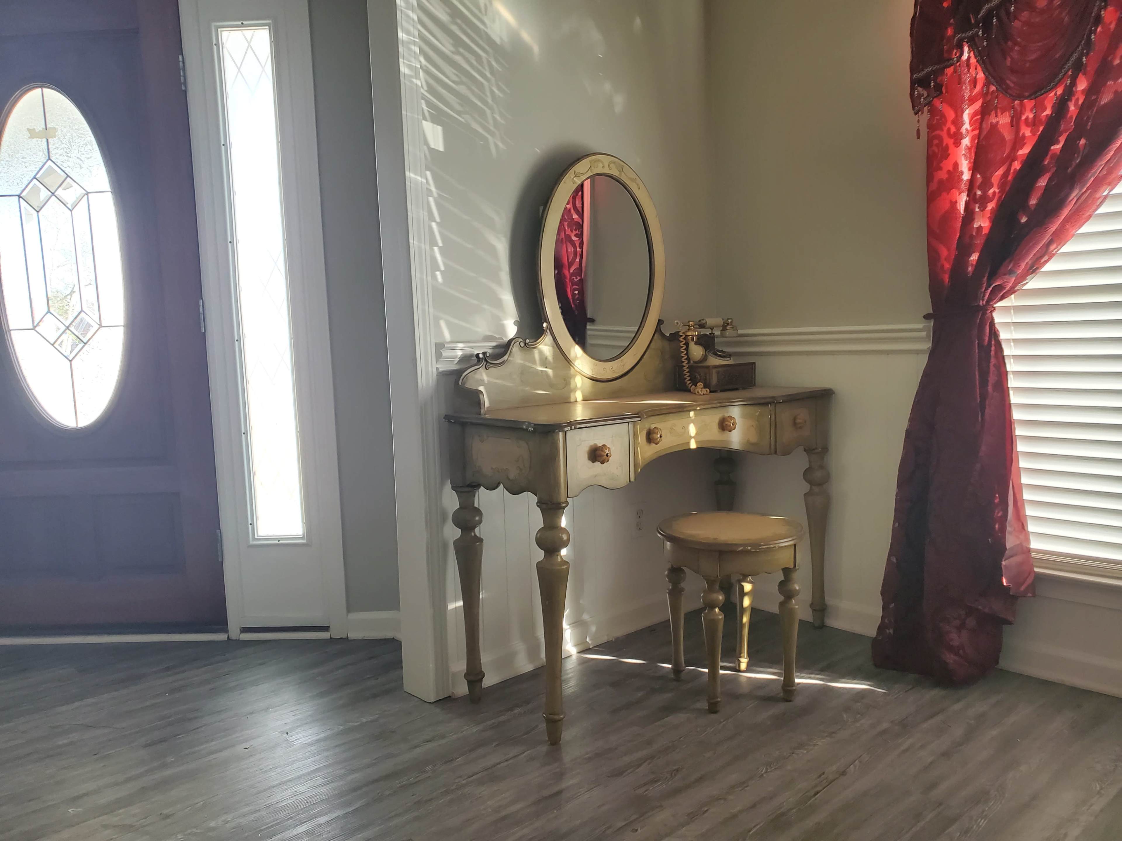 The scene features a vintage vanity table with a round mirror and a matching stool, positioned beside a doorway with decorative glass.
