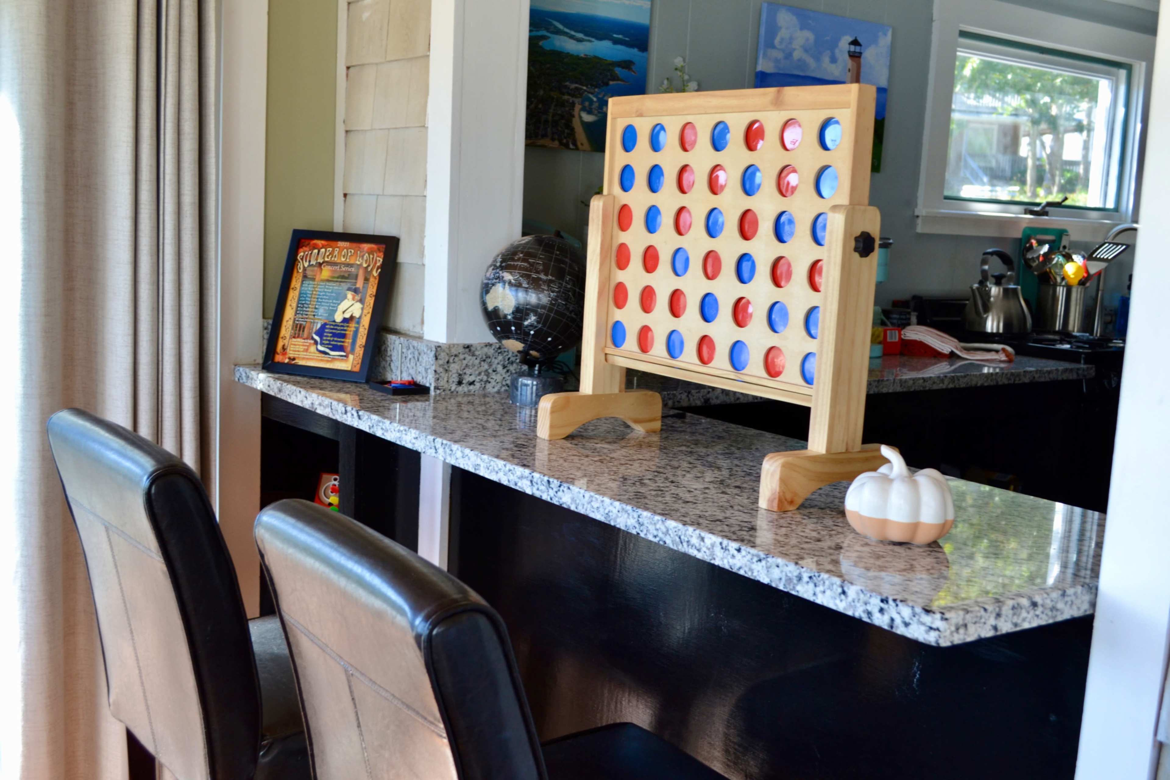 A Connect Four game is set up on a granite counter next to a framed picture and a globe, with two leather chairs in front.