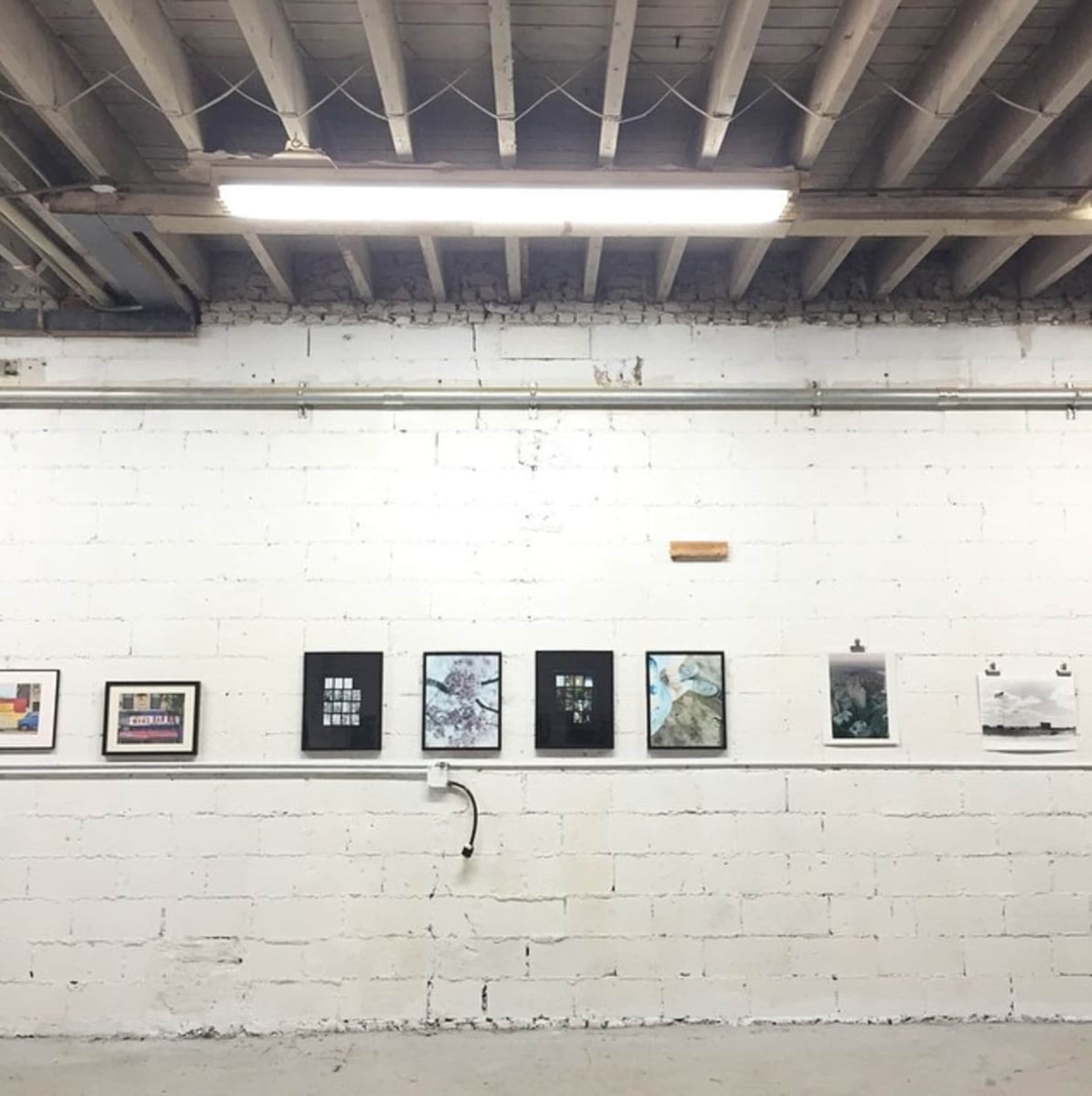 The image shows a minimalist gallery space with a white brick wall displaying several framed artworks and a few pieces on clipboards.