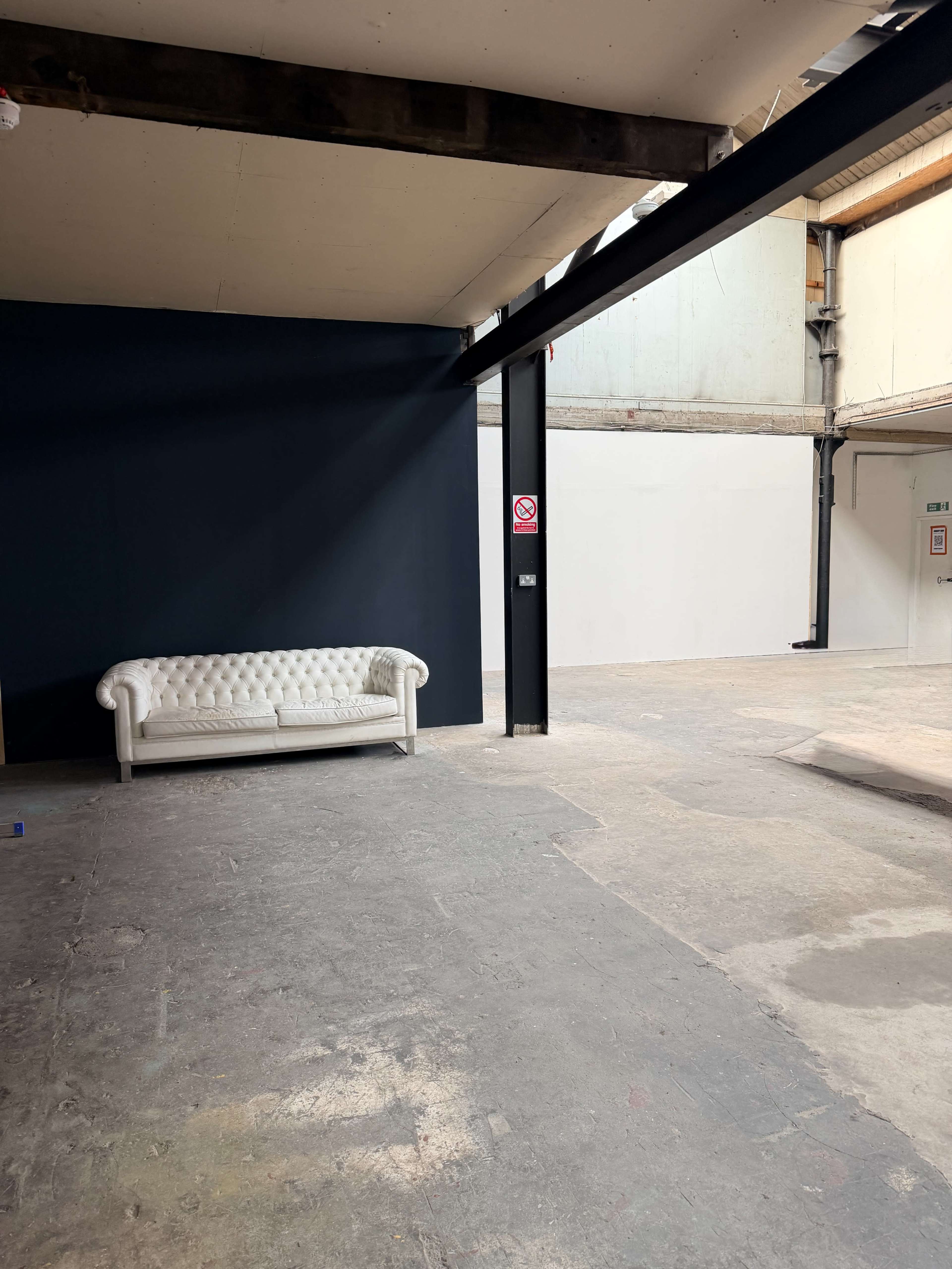 The image shows a spacious interior featuring a white chesterfield sofa against a dark blue wall, with exposed beams and concrete flooring.