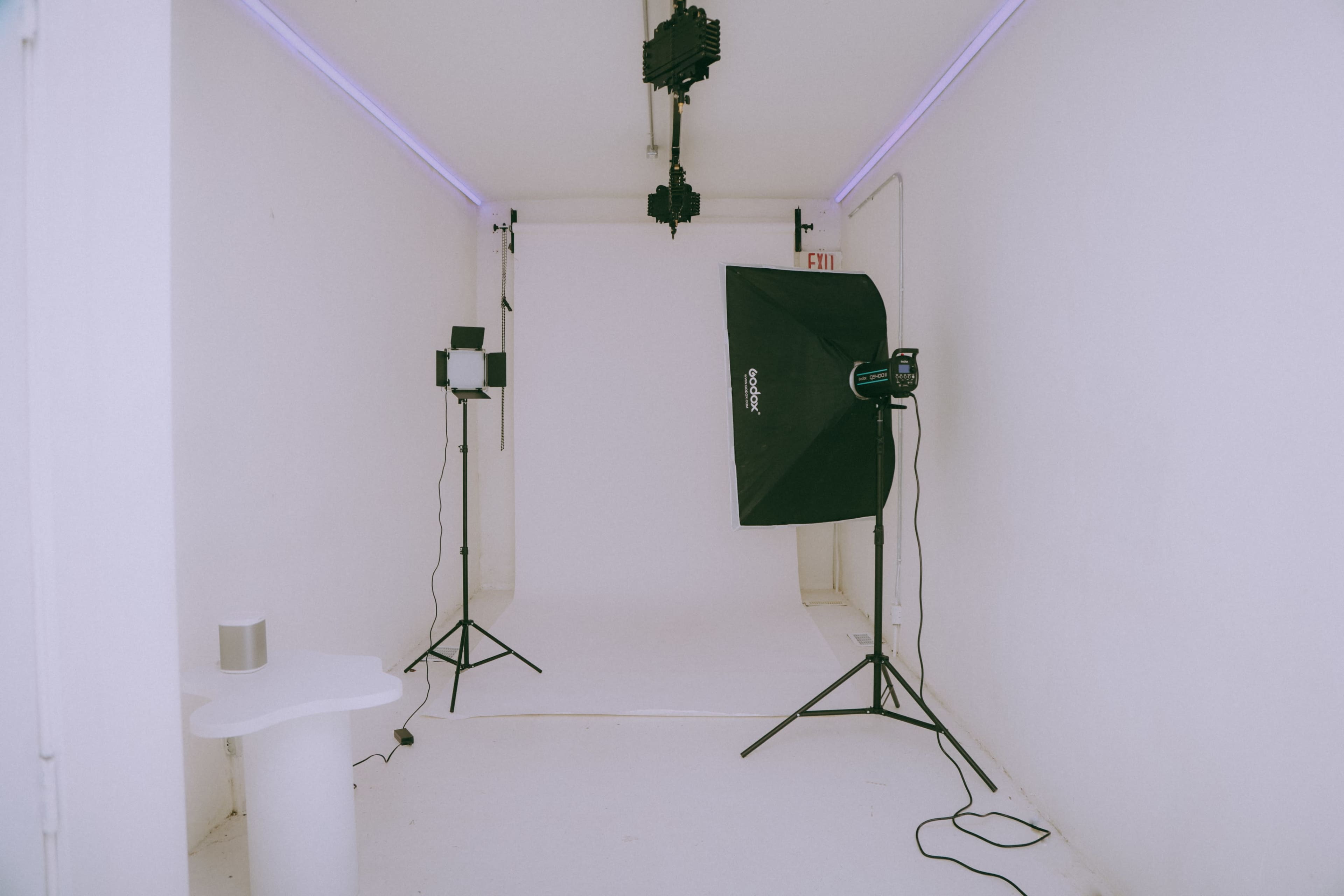 The image shows a minimalist photography studio featuring a white backdrop, lighting equipment on tripods, and a small table in the corner.
