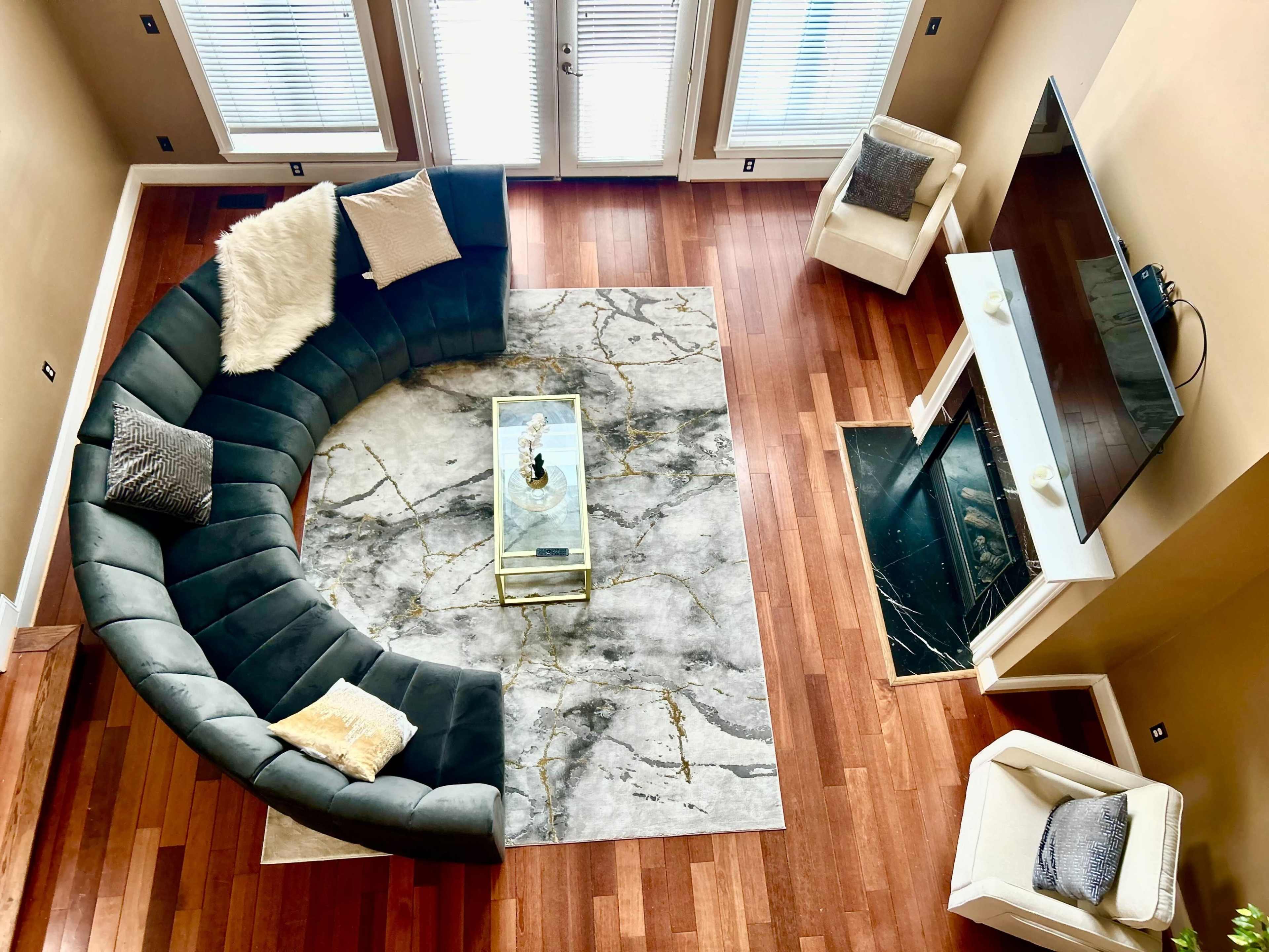 A modern living room features a curved gray sectional sofa, a marble-patterned rug, a glass coffee table, and a flat-screen TV above a fireplace.