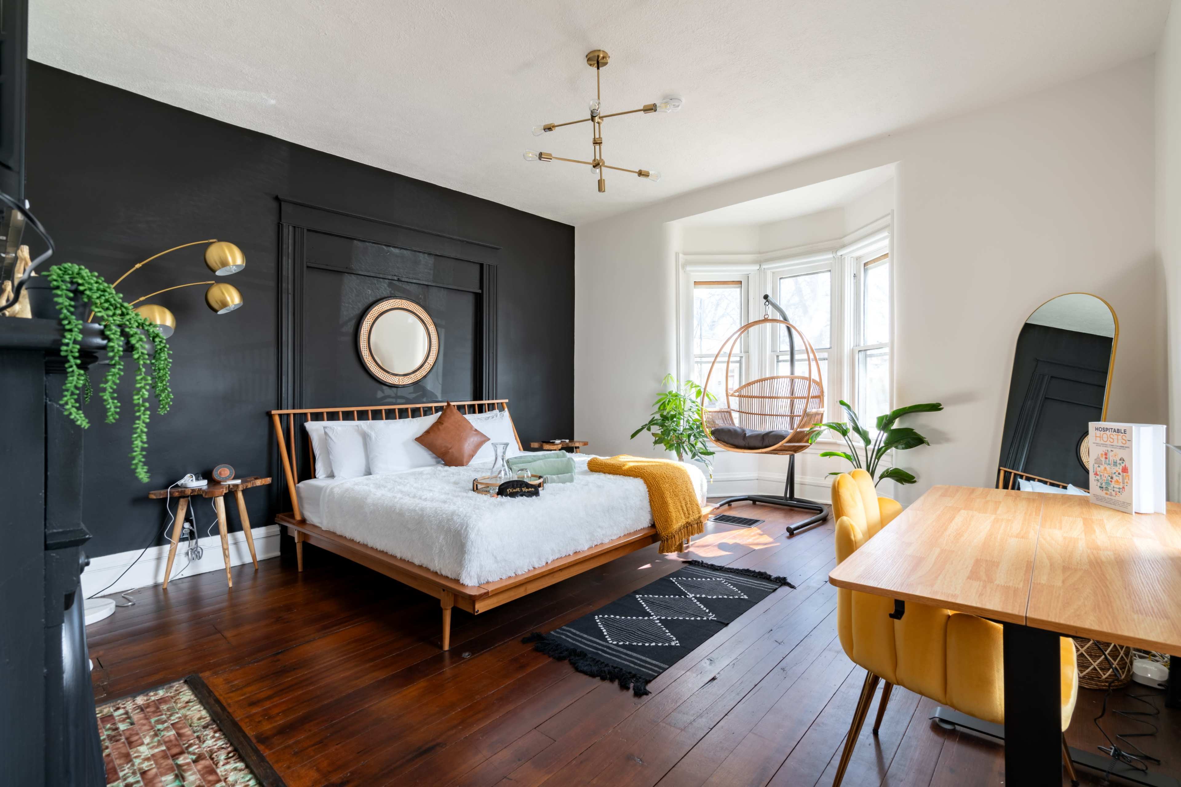 A modern bedroom featuring a white bed with decorative pillows, a hanging chair, a wooden desk with a yellow chair, and large windows allowing natural light.
