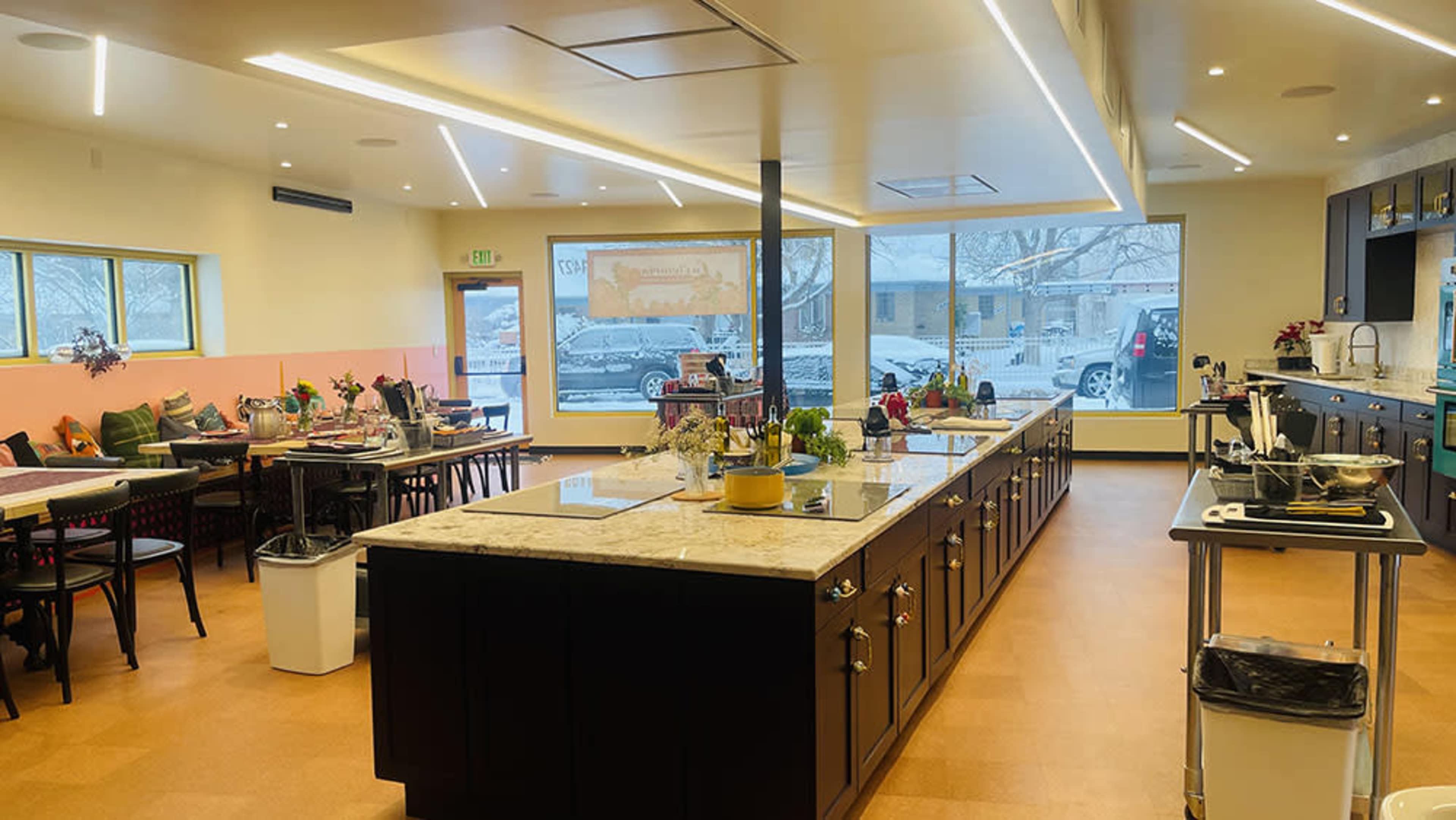 The image shows a modern kitchen and dining area with an island in the center, tables along the sides, and large windows letting in natural light.