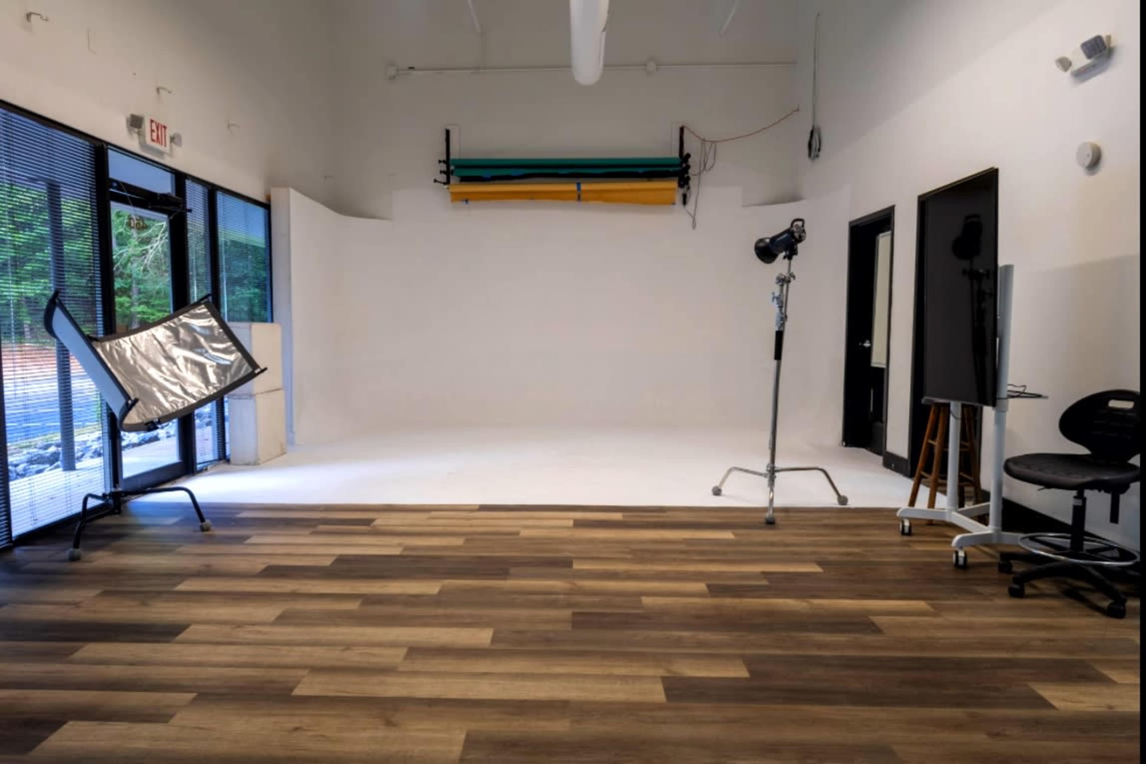 The image shows a photography studio with a plain white backdrop, wooden flooring, and various equipment including a reflector and a light stand.