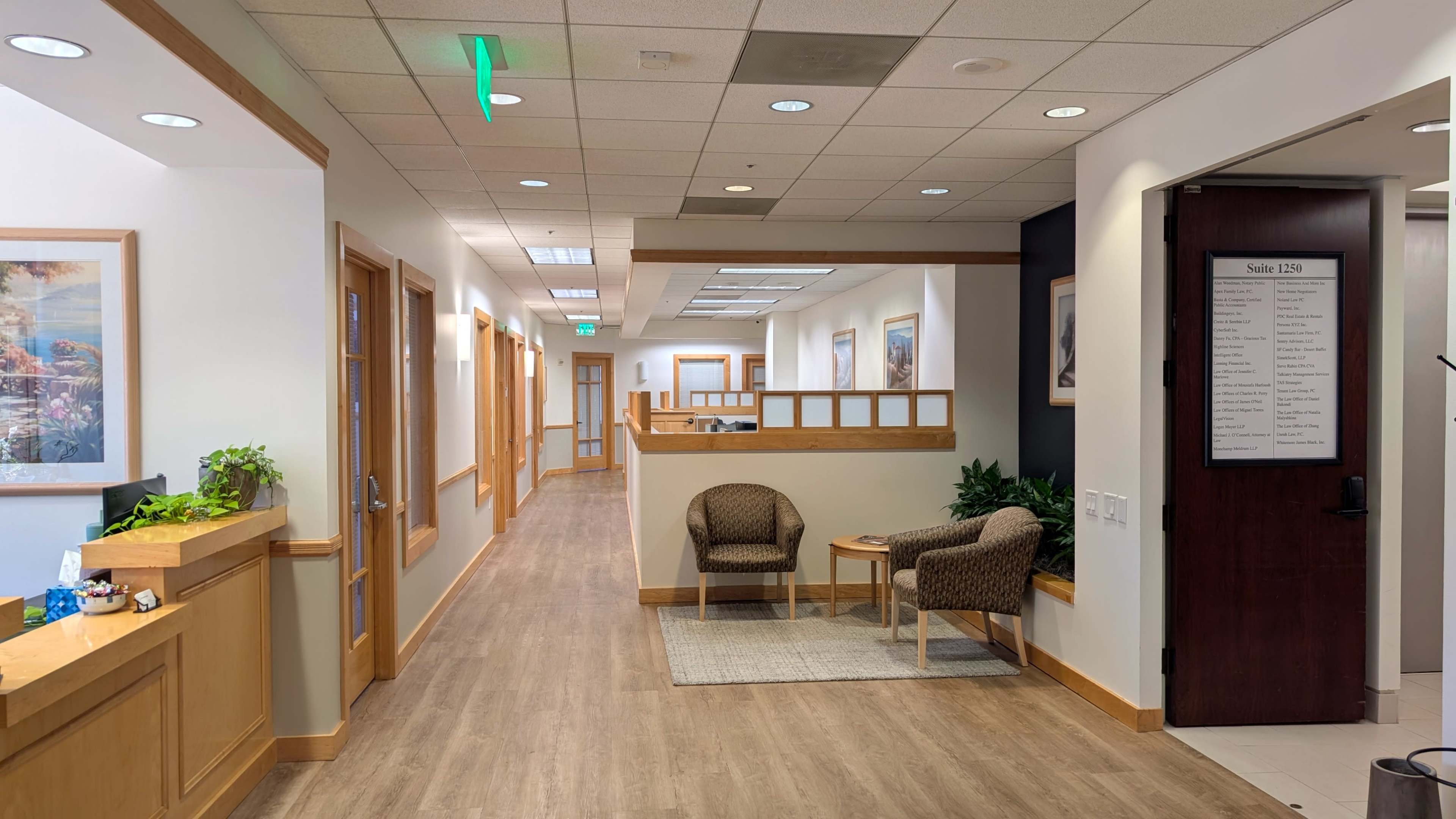 The image shows a brightly lit hallway in an office building with a seating area featuring two chairs and a small table, flanked by wooden doors leading to various rooms.